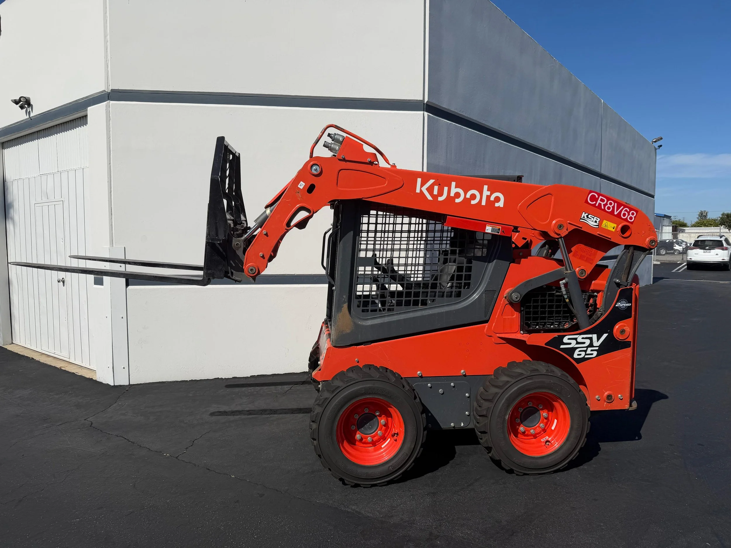 Orange Kubota skid-steer loader with black tires and front lift forks parked on black asphalt near a white and gray building under a clear blue sky. Skid steer available for rent in Santa Ana, CA