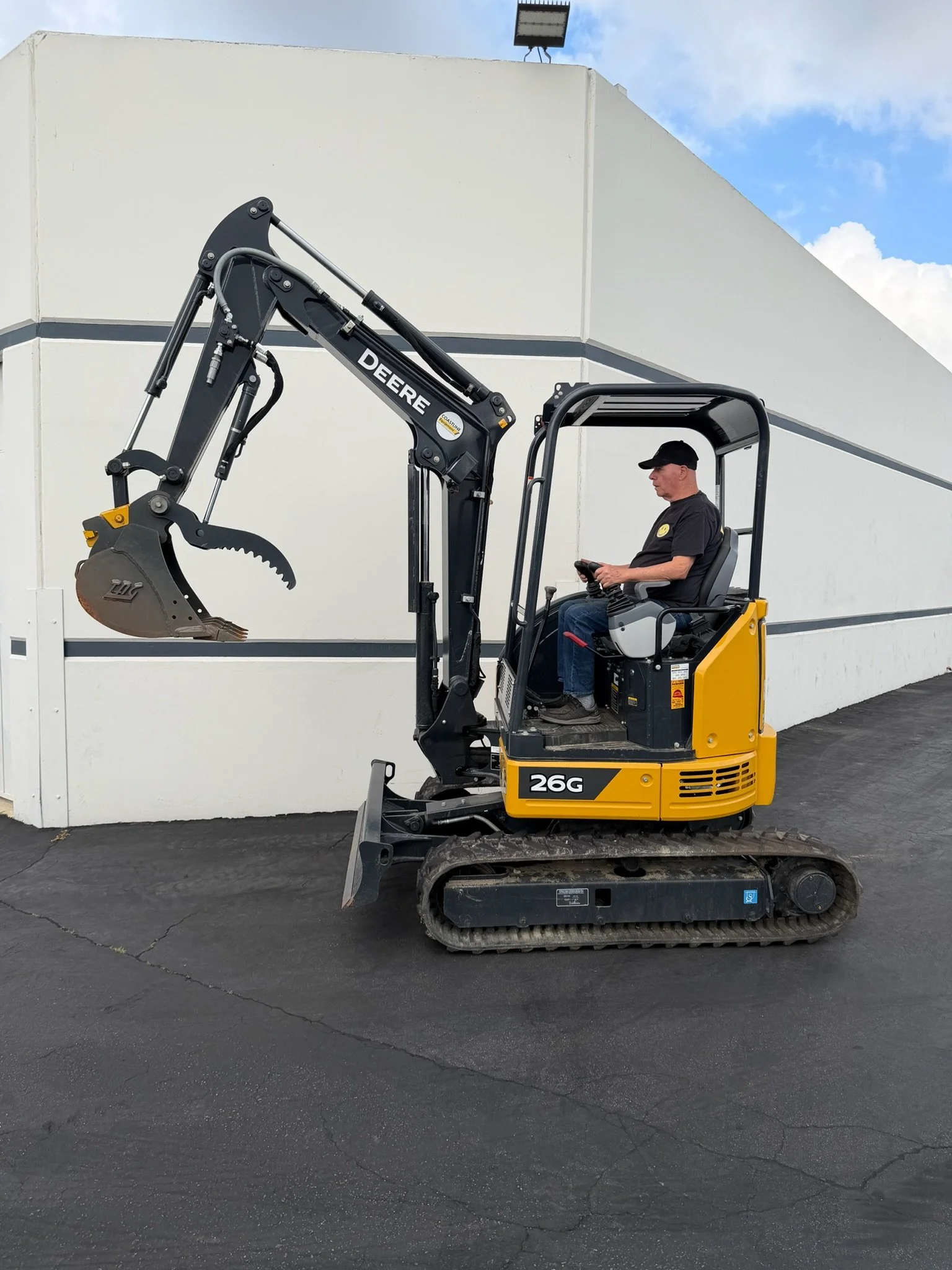 A man operating a small John Deere excavator on a paved surface near a white building with gray trim. Available for rent in Santa Ana, CA