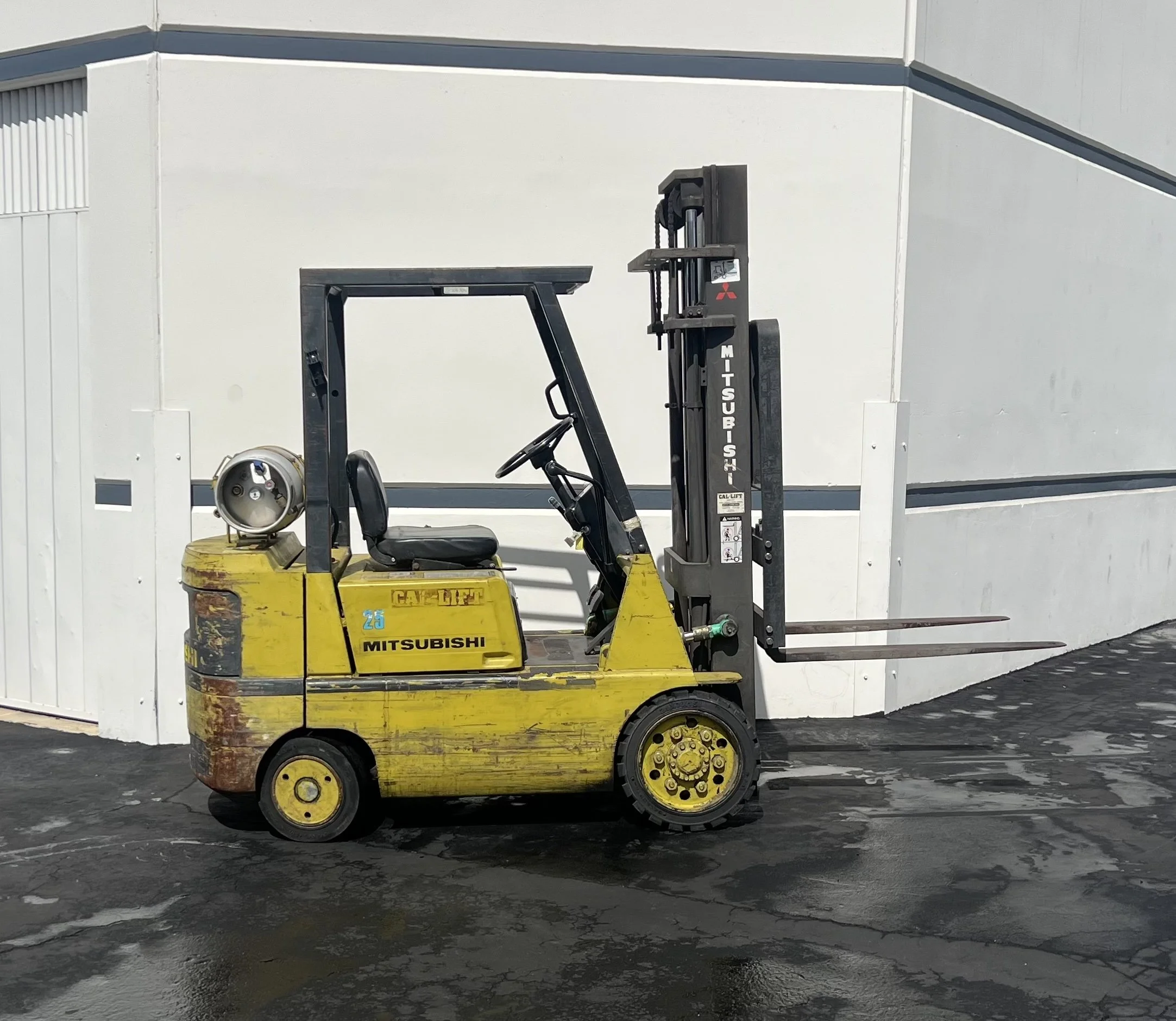 Yellow Mitsubishi forklift parked on a wet asphalt surface outside a white building.