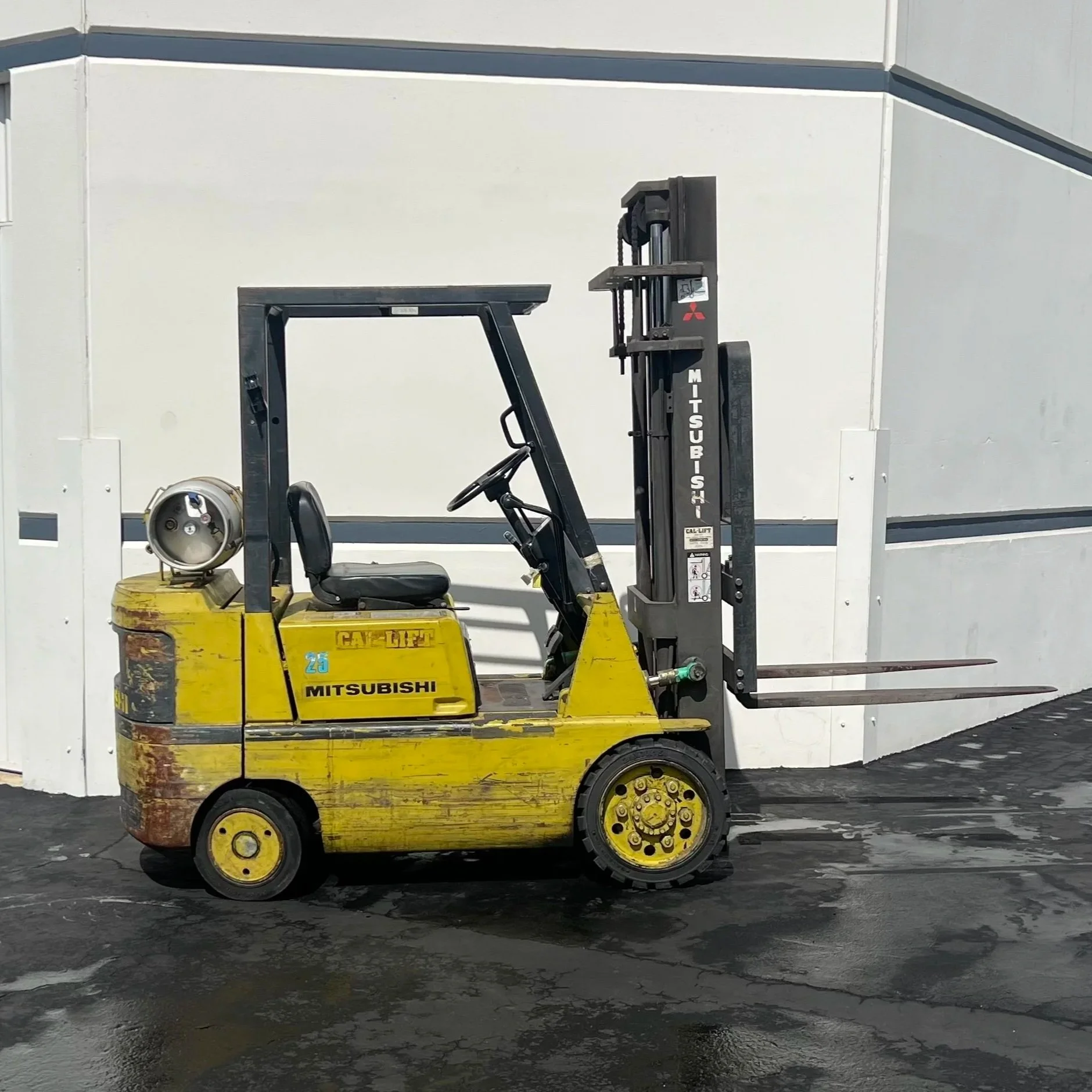 Yellow Mitsubishi forklift with some rust and wear, parked on a wet asphalt surface outside a white building. Forklift available for rent in Santa Ana, CA