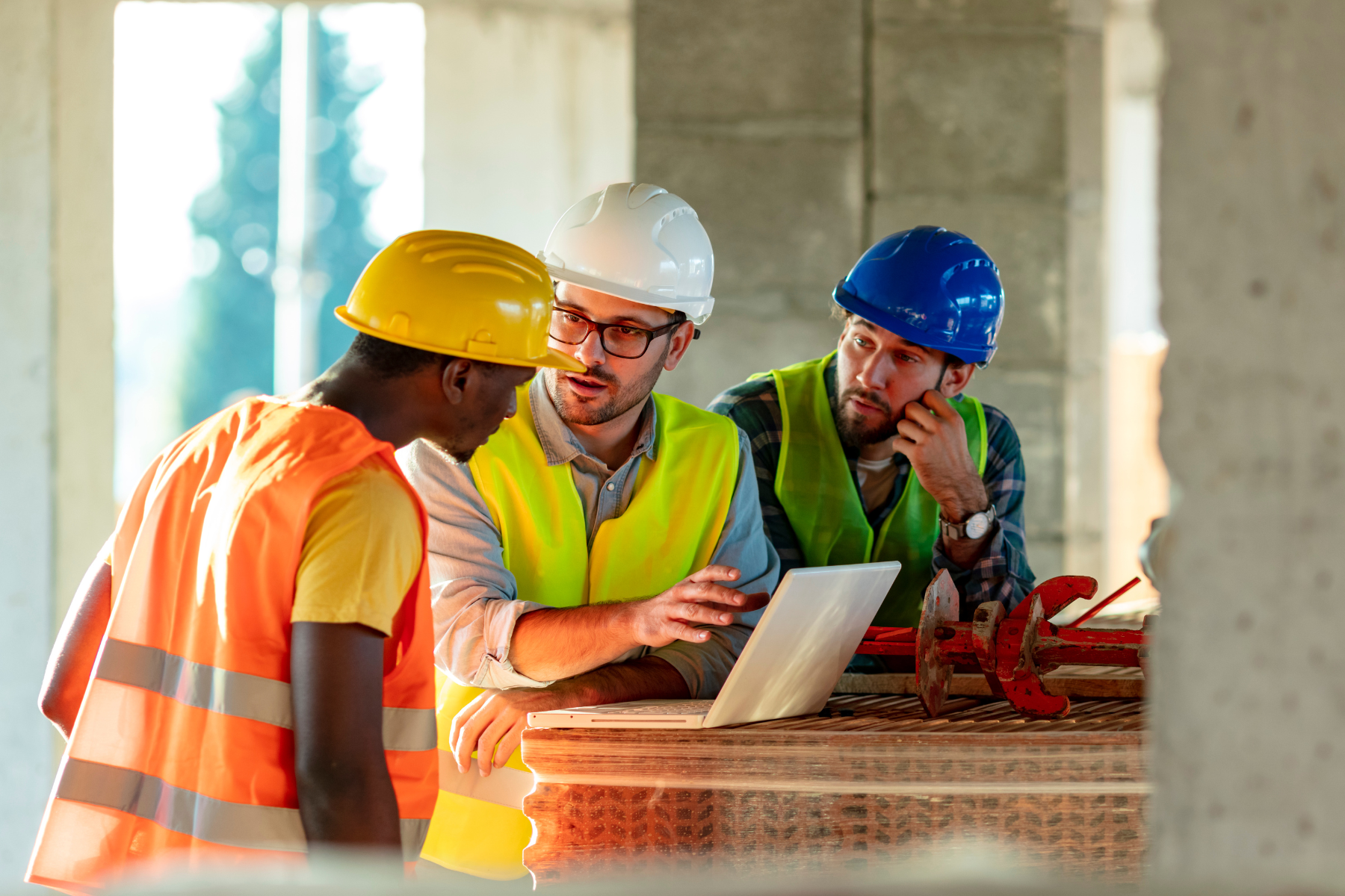 Three construction workers, two wearing blue and white hard hats and one wearing a yellow hard hat, are discussing plans at a construction site with a laptop on the wooden table. Construction equipment rentals in Santa Ana, CA