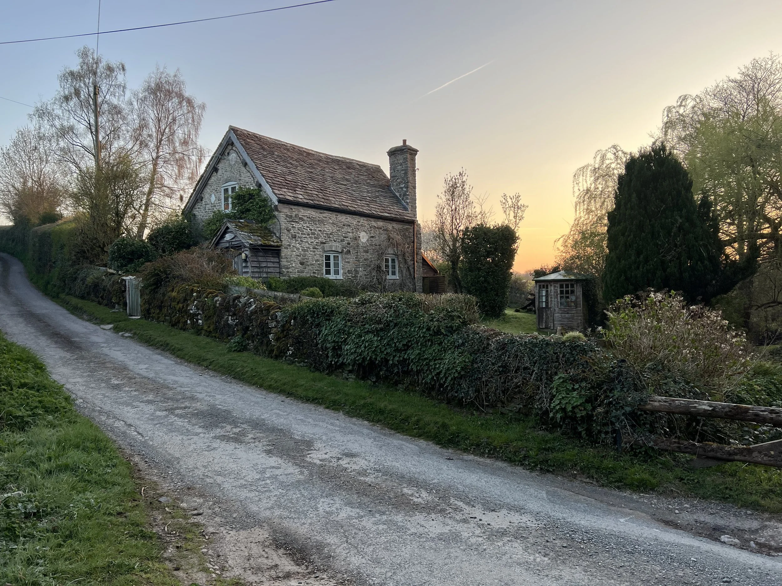 A rural stone house with a tiled roof and a chimney, surrounded by trees and bushes, at sunset. A dirt road runs in front of the house.