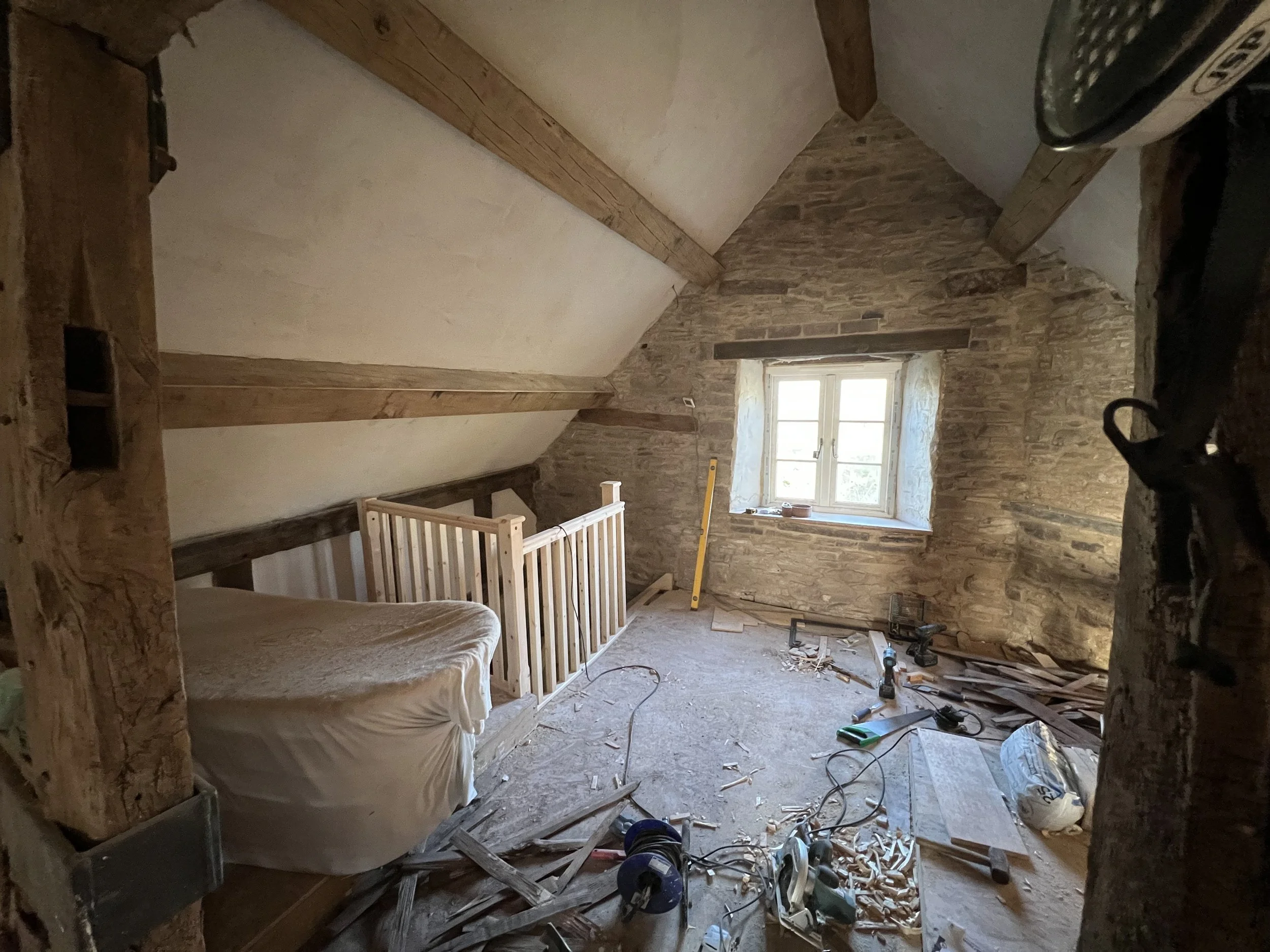 A room under renovation with exposed brick walls, a sloped ceiling with wooden beams, a small window letting in natural light, and construction tools and debris on the floor.