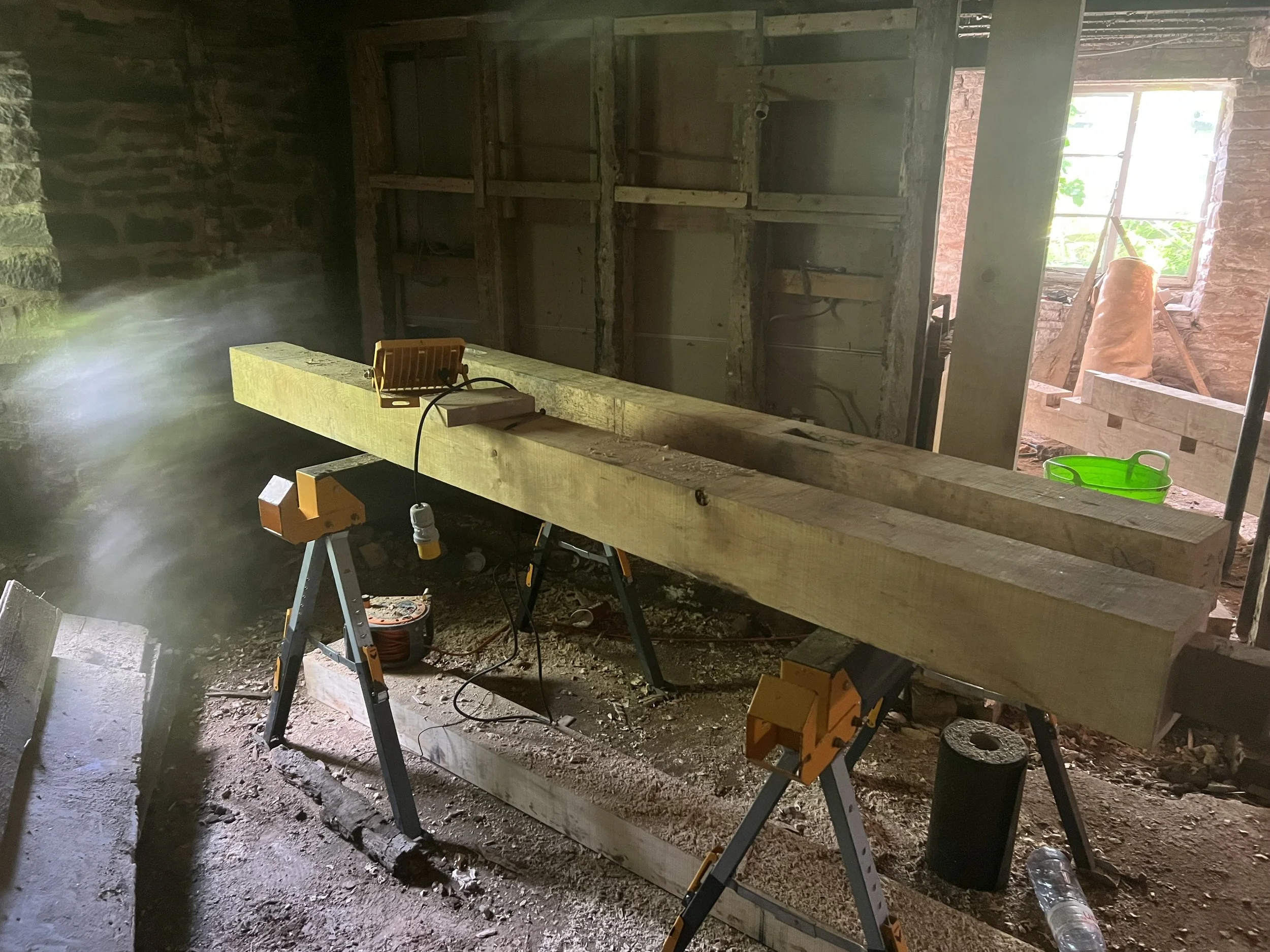 Construction site with a large wooden beam on sawhorses, window in the background, and construction tools around.