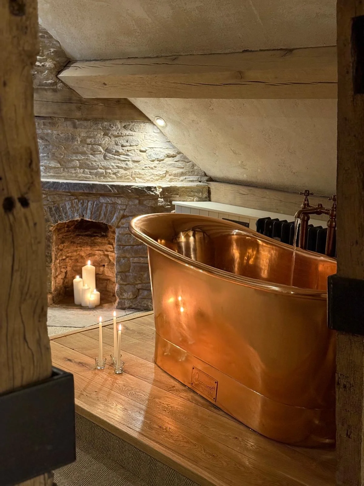 A copper bathtub in a cozy rustic room with a stone fireplace, candles on the floor, and soft lighting.