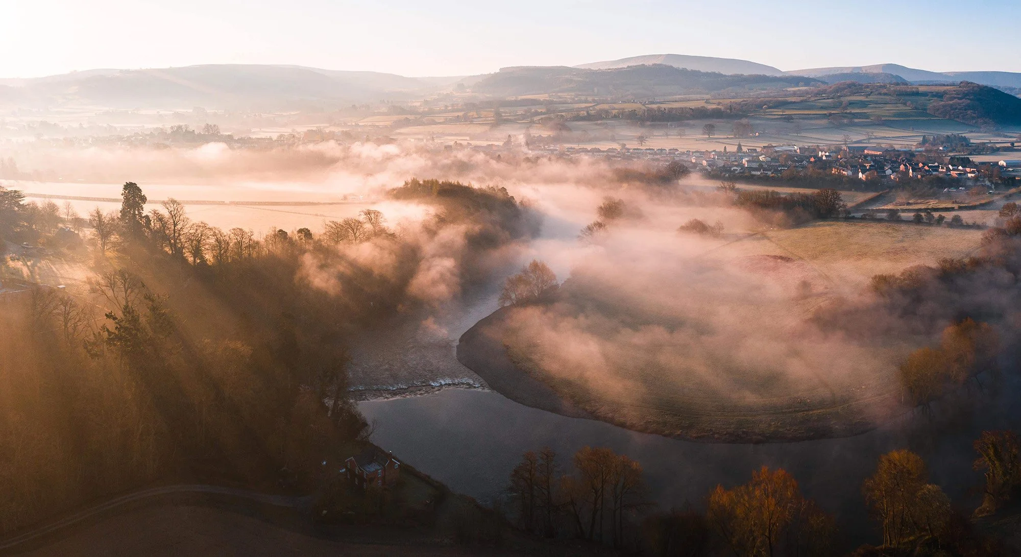 A misty river winding through a valley with fog rising from the water, surrounded by leafless trees and rolling hills under a clear sky, early morning sunlight casting long shadows.