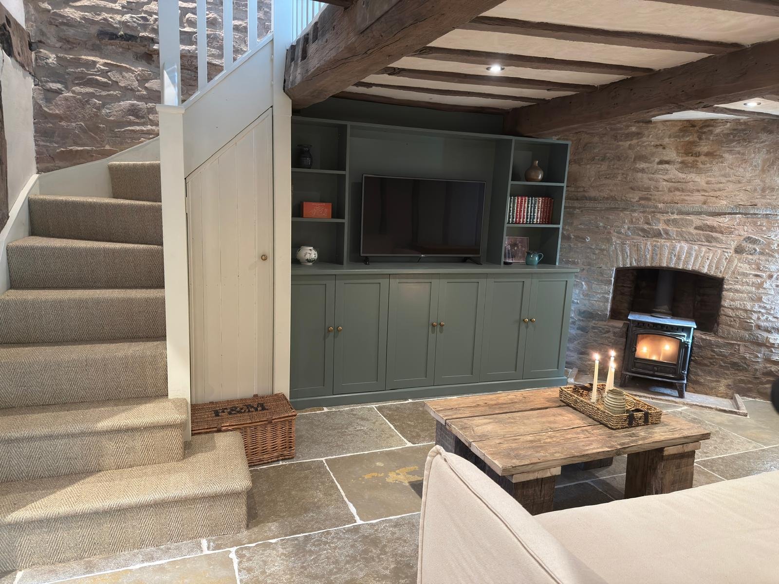Living room with stone walls, a staircase with beige carpeting, a green built-in shelf with TV, a rustic wooden coffee table with candles, and a small stove in the fireplace.