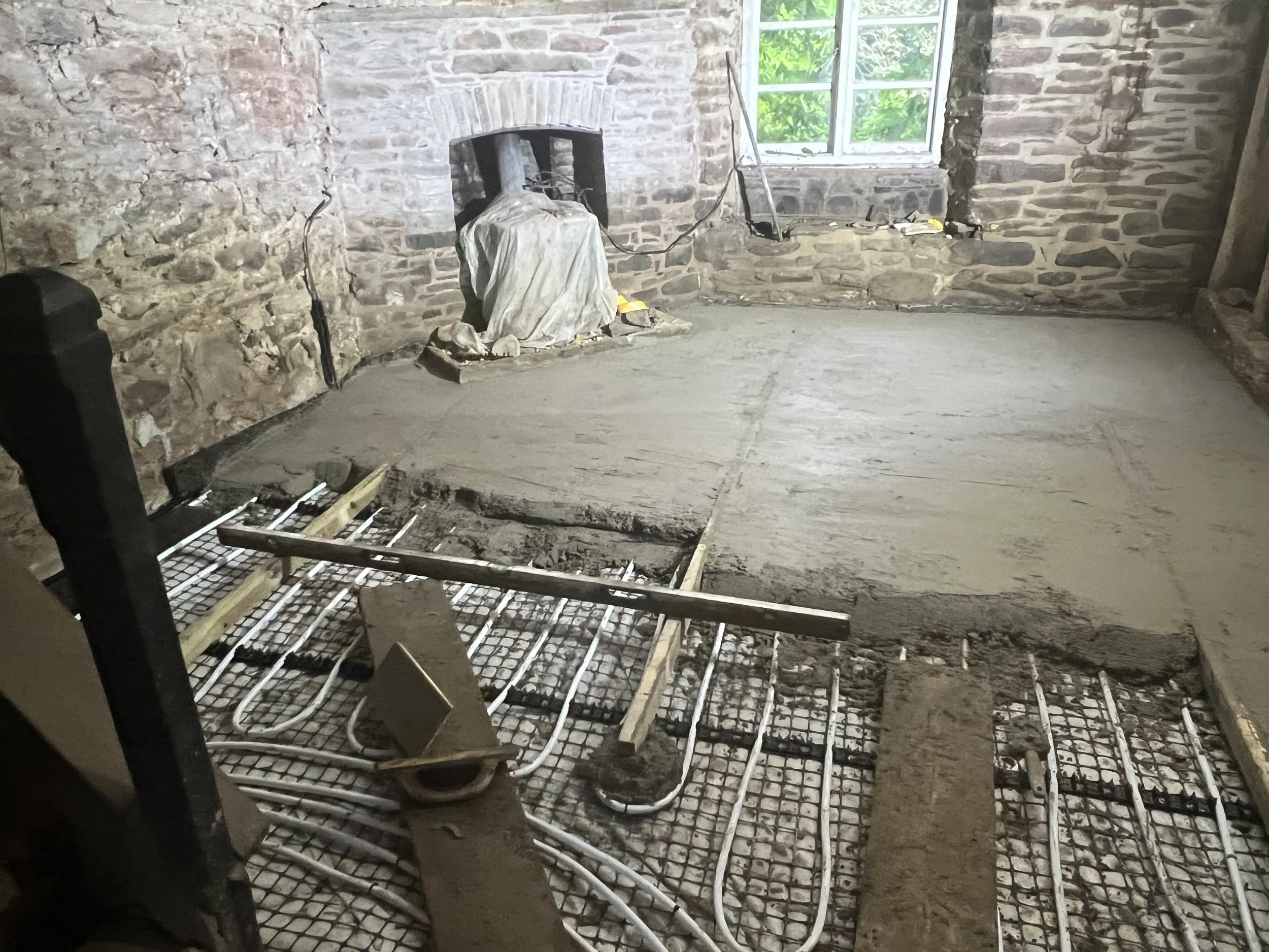 Interior of a room under renovation with a concrete floor being poured, exposed brick walls, a window with green trees outside, and construction tools and piping visible.