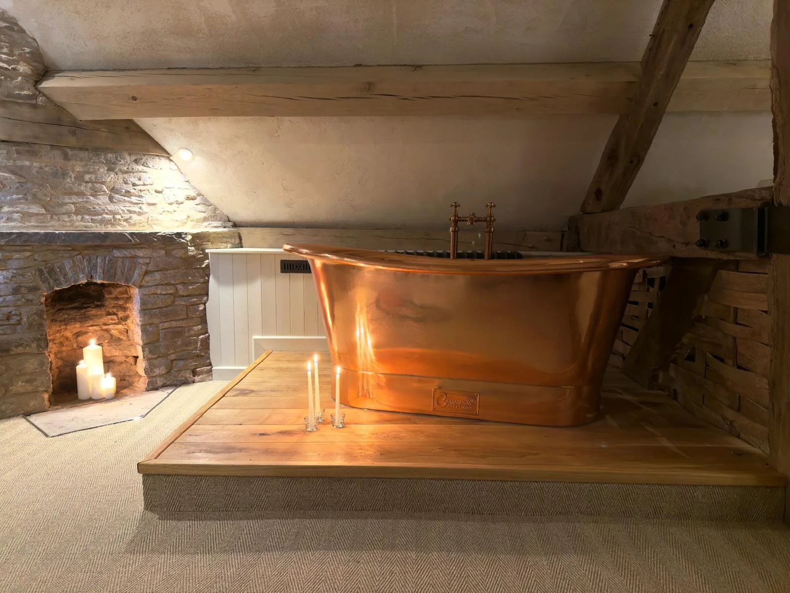 A copper bathtub on a wooden platform in a rustic bathroom with exposed brick and wooden beams, decorated with candles.