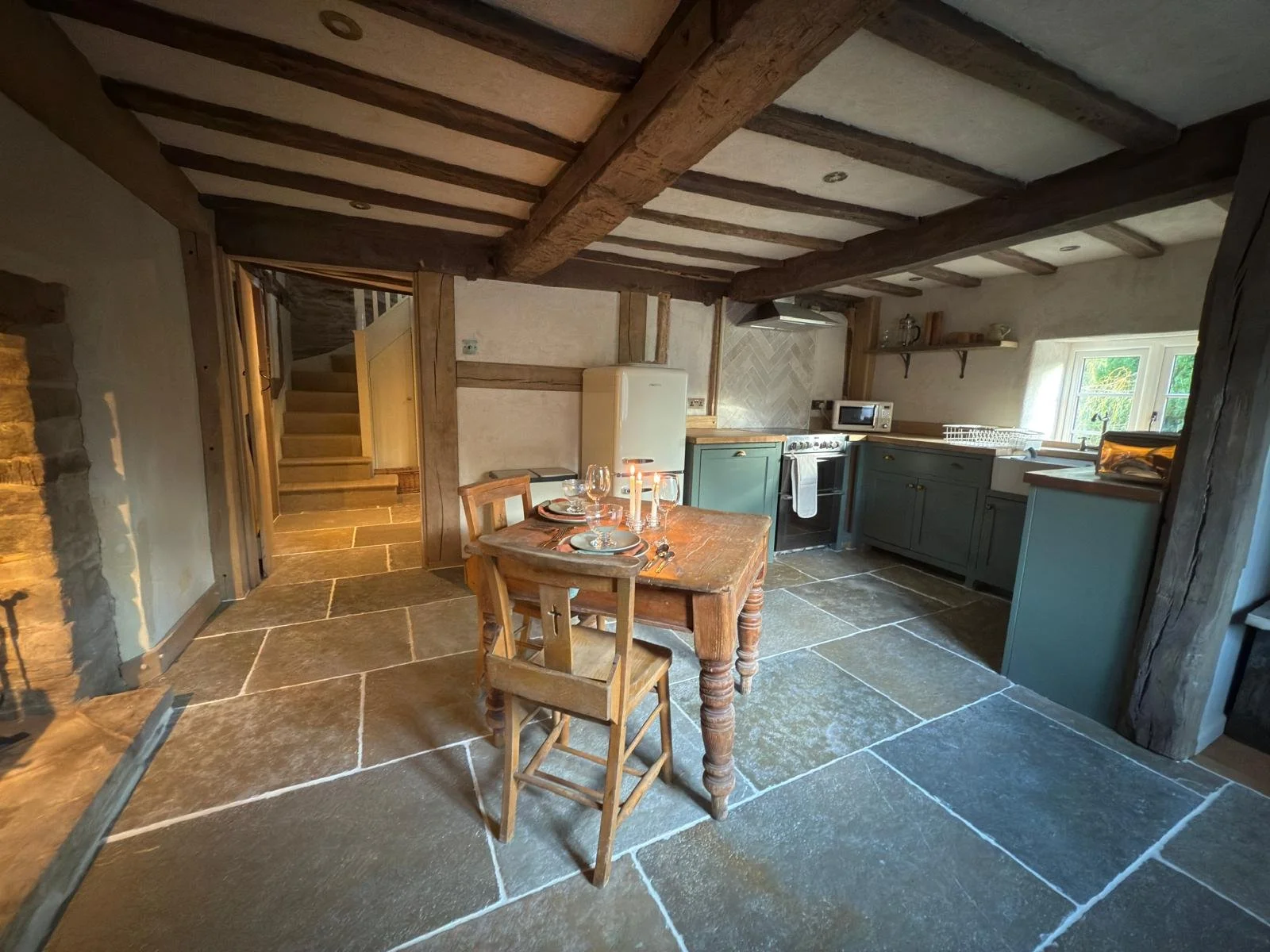 Kitchen with stone floor, exposed wooden beams, green cabinets, a white fridge, stove, microwave, window, and a wooden dining table set for two with candles.