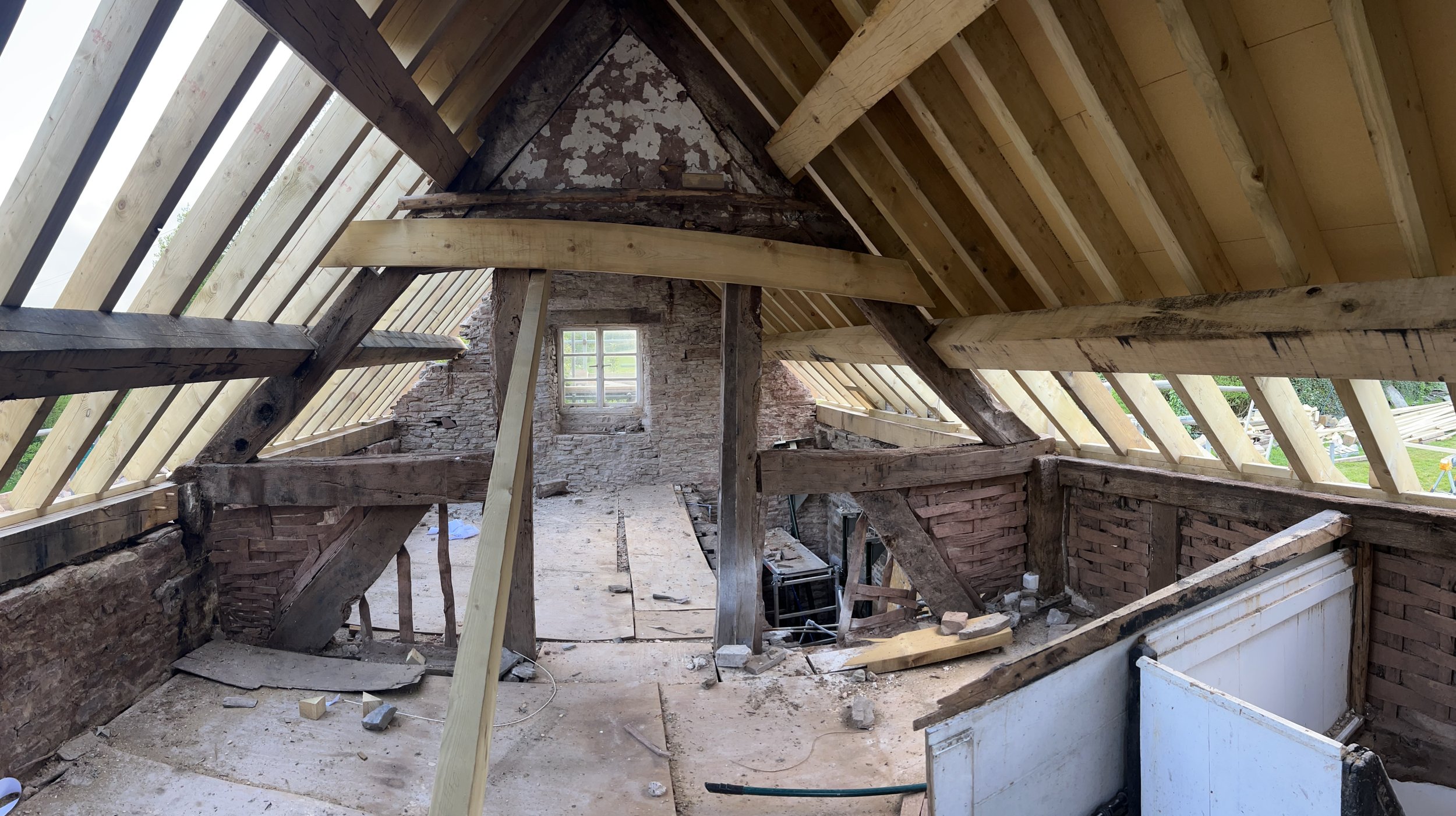 Attic under renovation showing exposed brick walls, wooden beams, and new roof framing with skylights. There is construction debris and tools on the floor.
