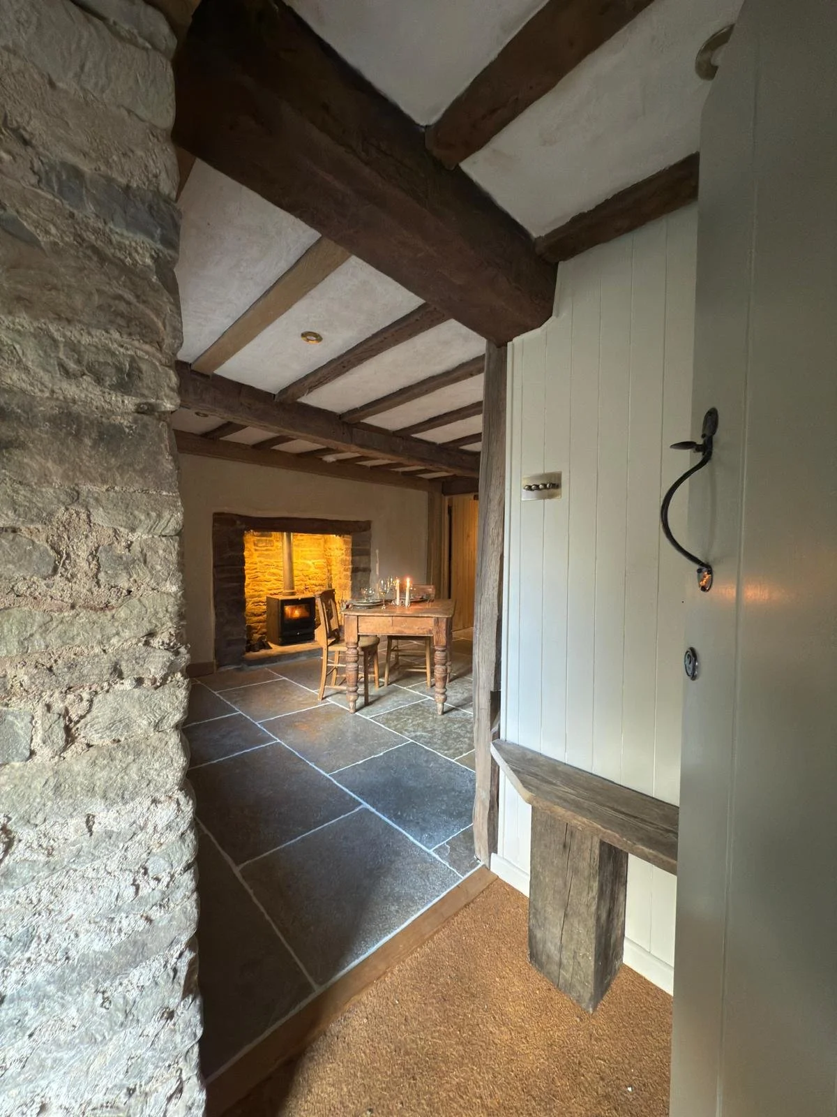 View of a cozy rustic living room with stone and wood accents, a stone fireplace, and a small table with chairs, seen through an entryway with white painted wood paneling.