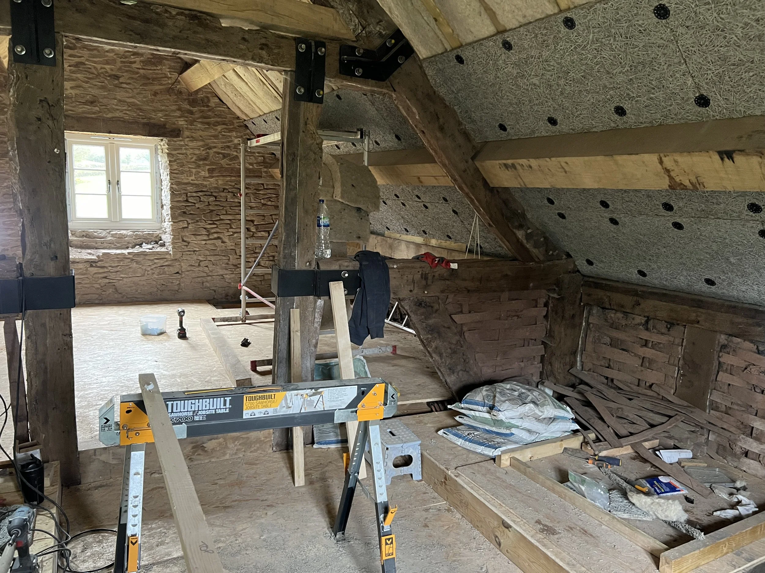 Interior of an attic under renovation with exposed wooden beams and brick walls, construction tools, and materials scattered around.