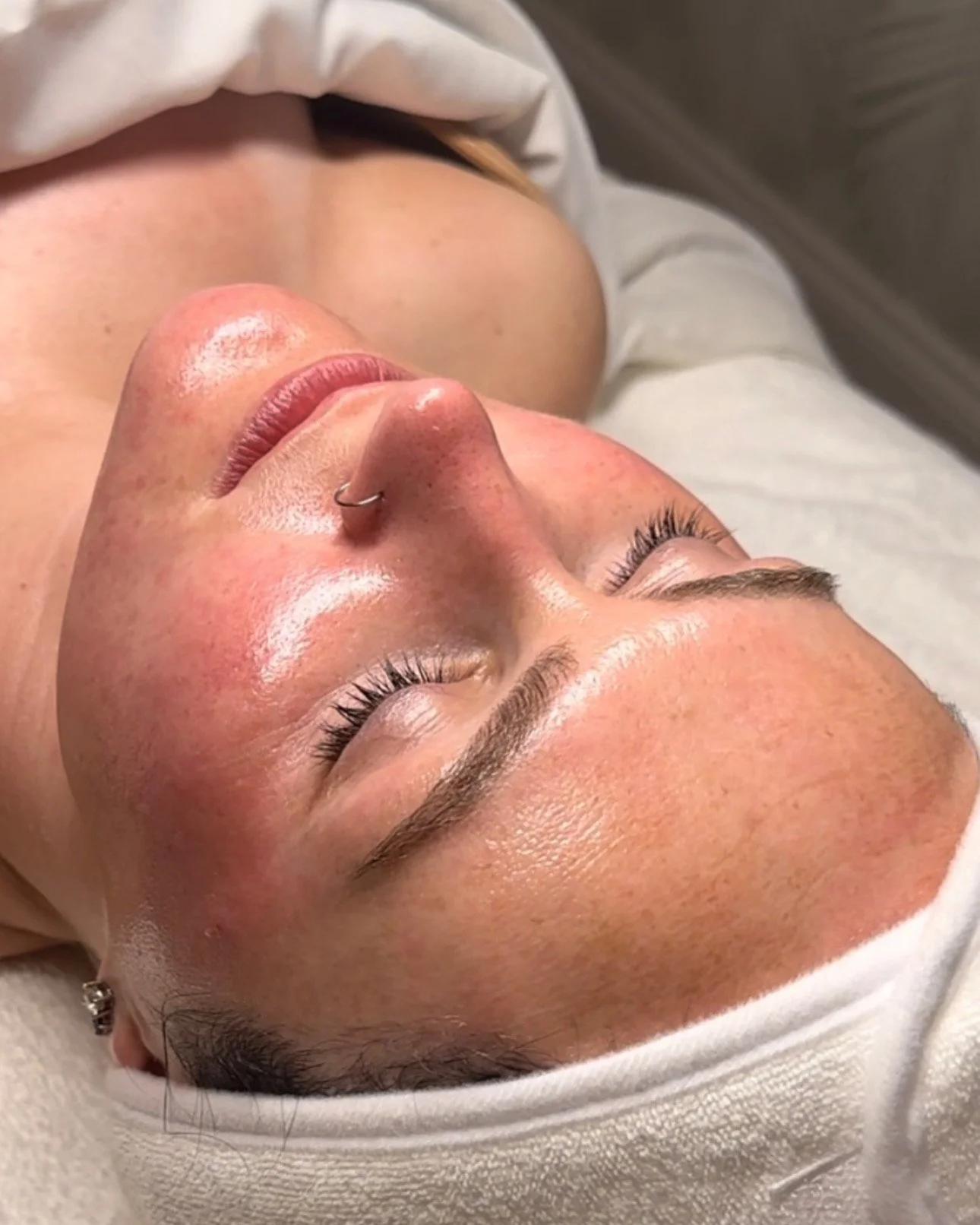 Close-up of a woman lying down with her eyes closed, showing her face and part of her shoulder, with natural lighting and a neutral background.