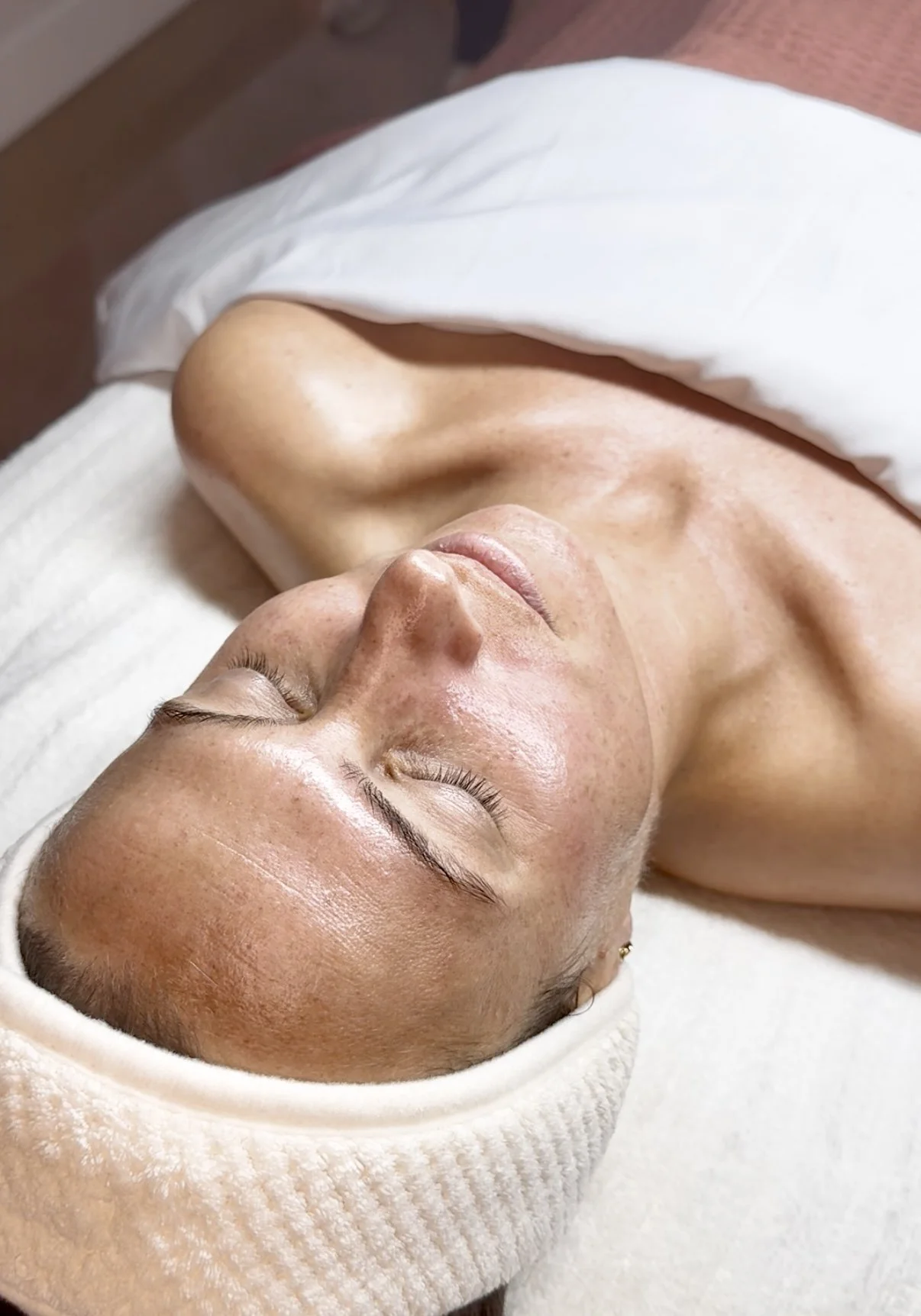 A woman lying down with her eyes closed, receiving a facial treatment, wrapped in a towel and wearing a headband.