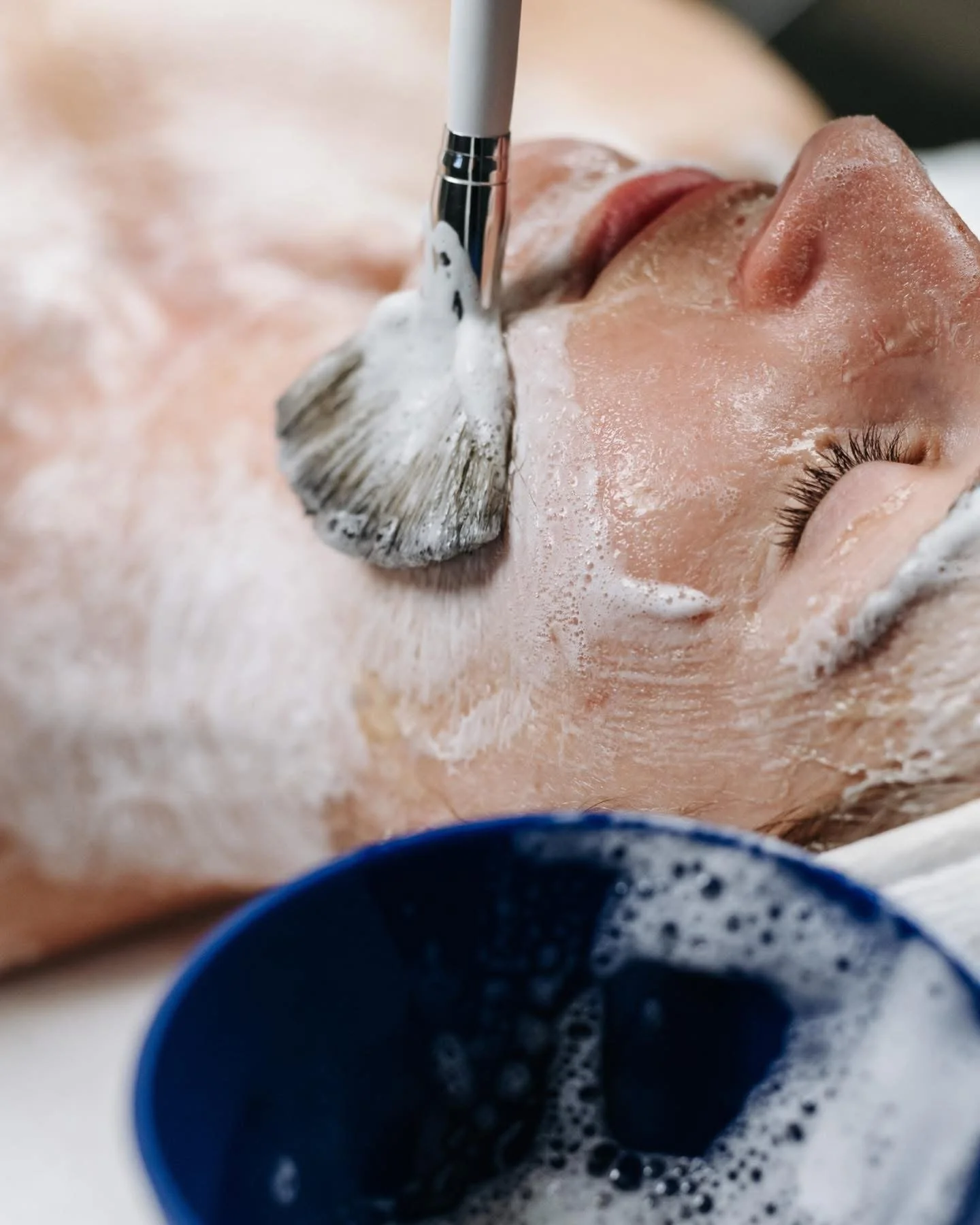 Close-up of a person receiving a facial cleansing treatment with a brush, with soap suds on their face and a blue basin of soapy water nearby.