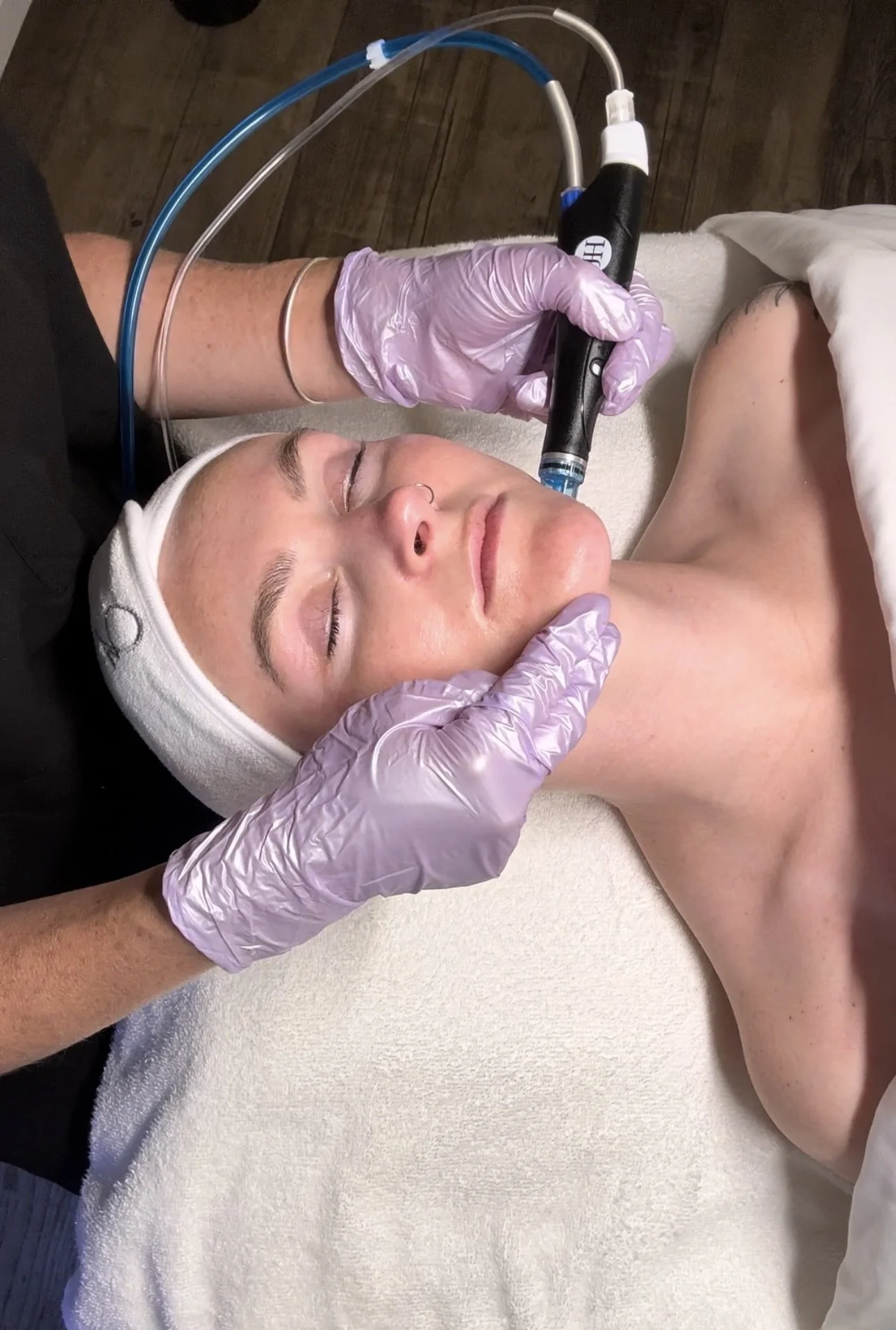 A woman lying face up with closed eyes and a white headband, receiving a facial treatment with a handheld device from a professional wearing purple gloves.