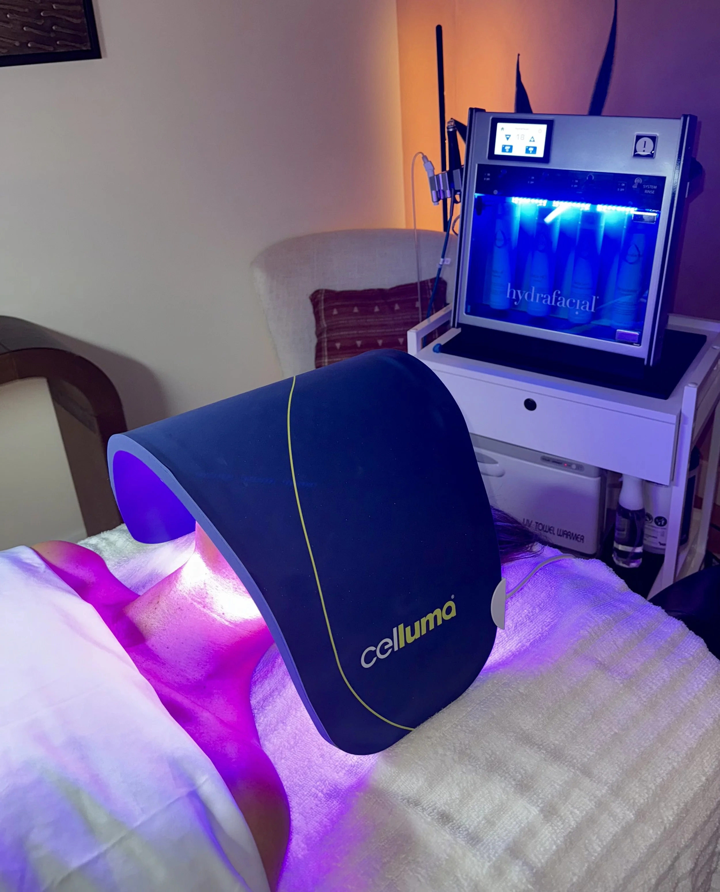 A person undergoing a hydrafacial treatment in a spa or clinic setting, with a large LED device emitting purple and pink lights positioned over their face, and a HydraFacial machine with skincare products in the background.