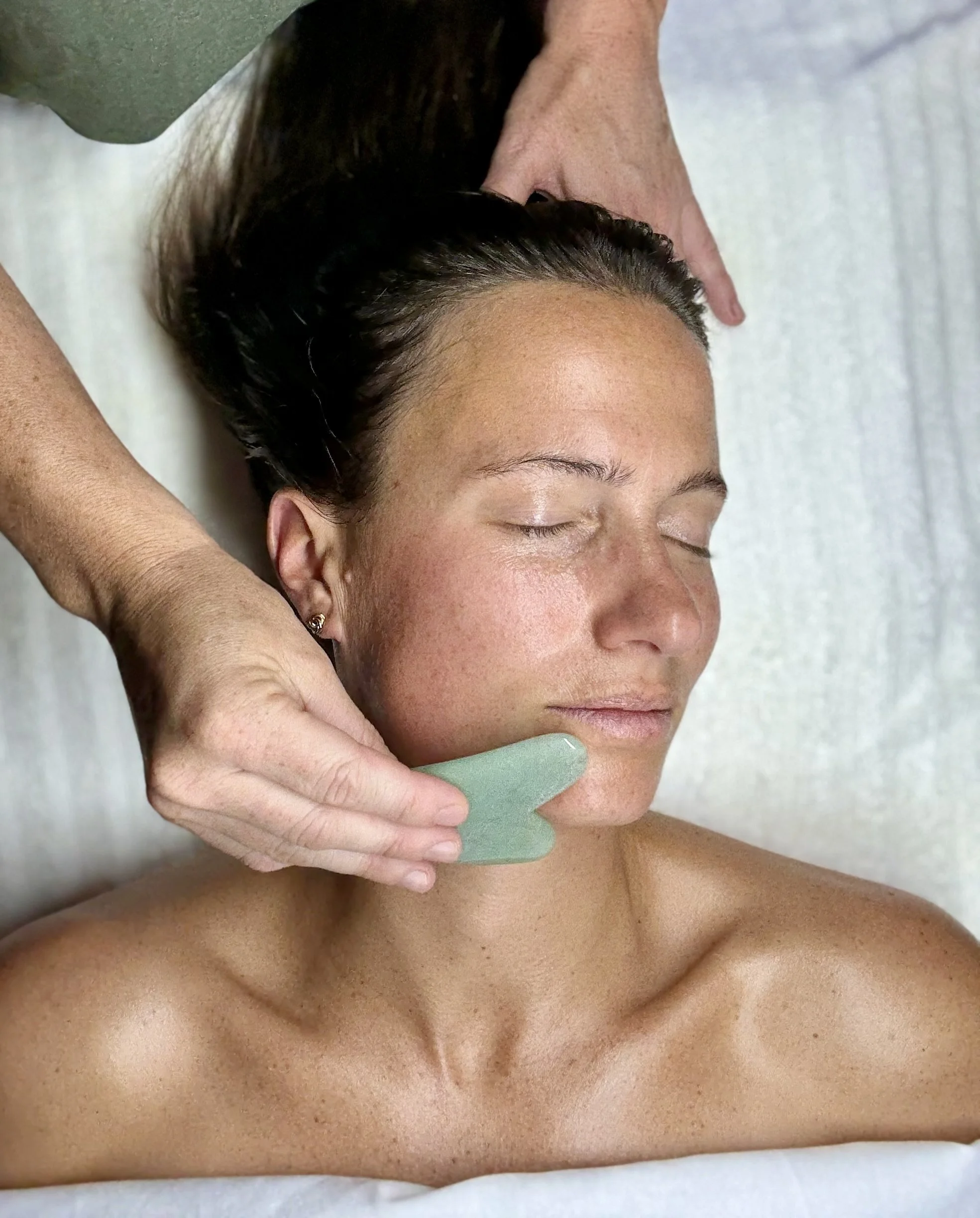 A woman receiving a facial massage or skincare treatment with a green gua sha tool on her cheek, with her eyes closed as she relaxes.