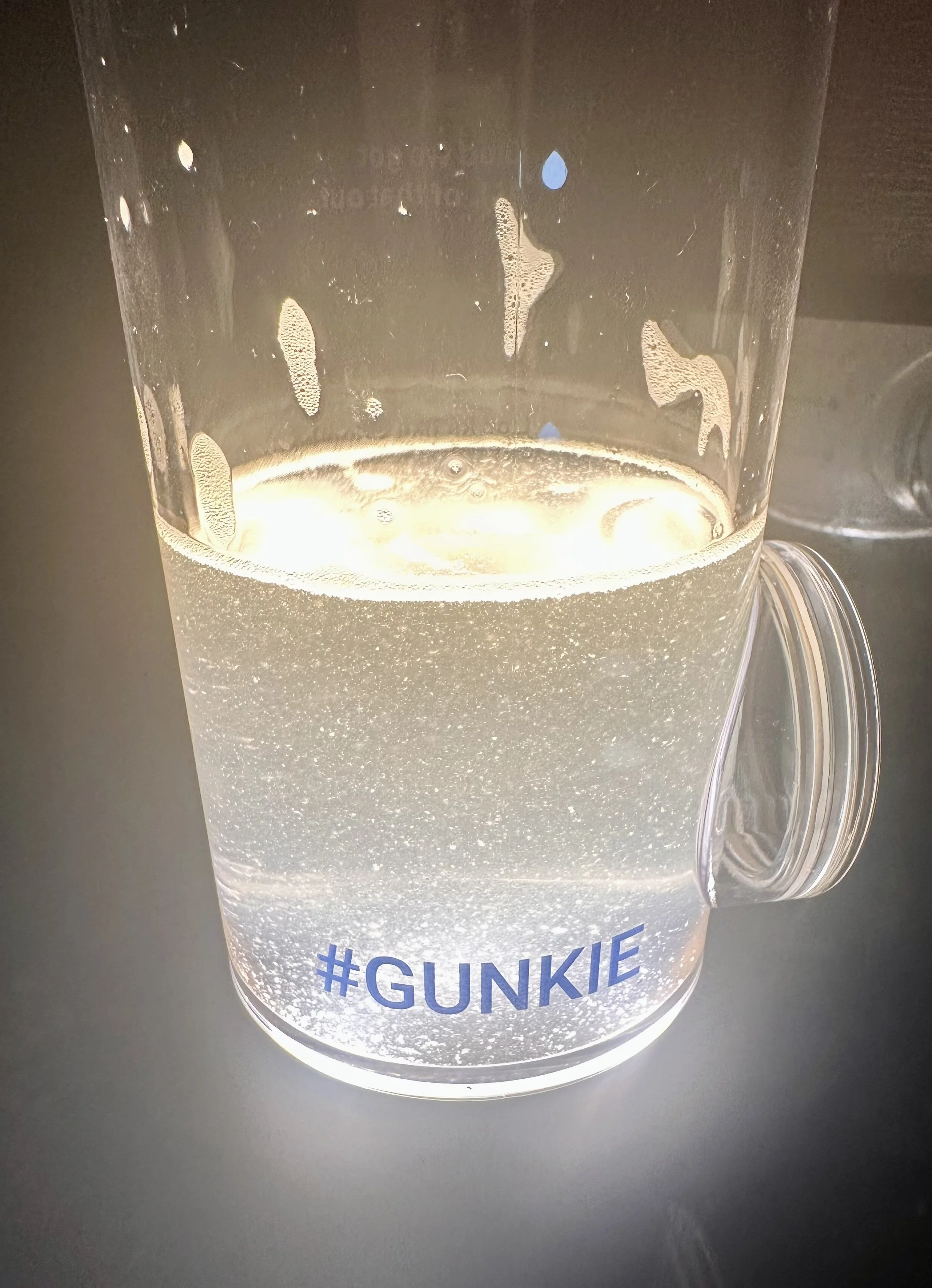 A clear plastic mug with the hashtag #GUNKIE printed on it, containing a partially filled, slightly fizzy liquid that appears to be beer, sitting on a dark surface.