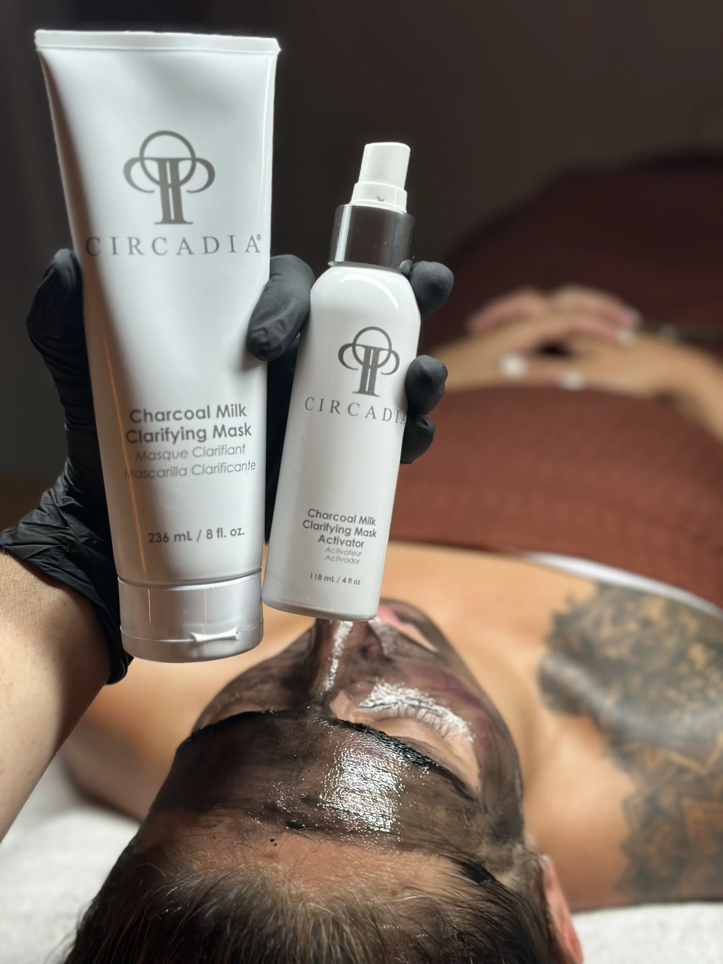 A person receiving a charcoal milk clarifying facial mask at a spa or skincare clinic, with products labeled 'Circadia' in their hand.