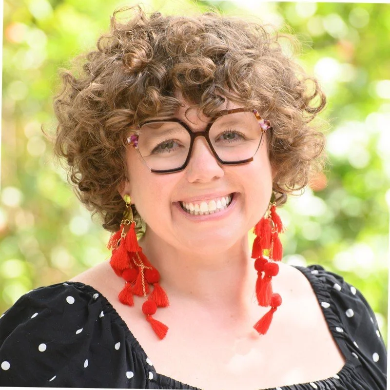 Smiling woman with curly hair, glasses, and red tassel earrings outdoors.