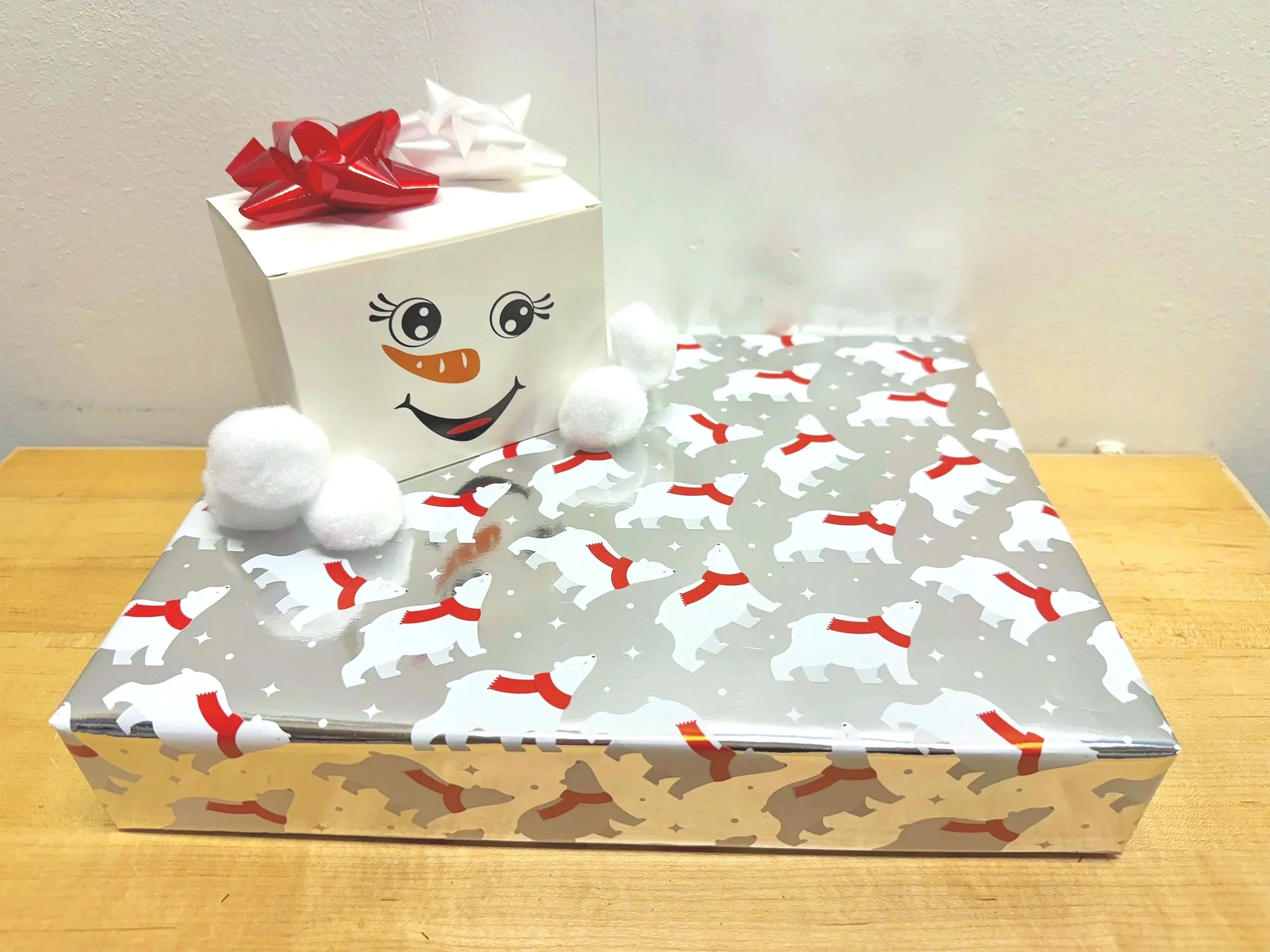 Christmas gift box with a snowman face, decorated with a red bow and white pom-poms, placed on a table with Christmas-themed wrapping paper featuring dogs wearing red scarves.