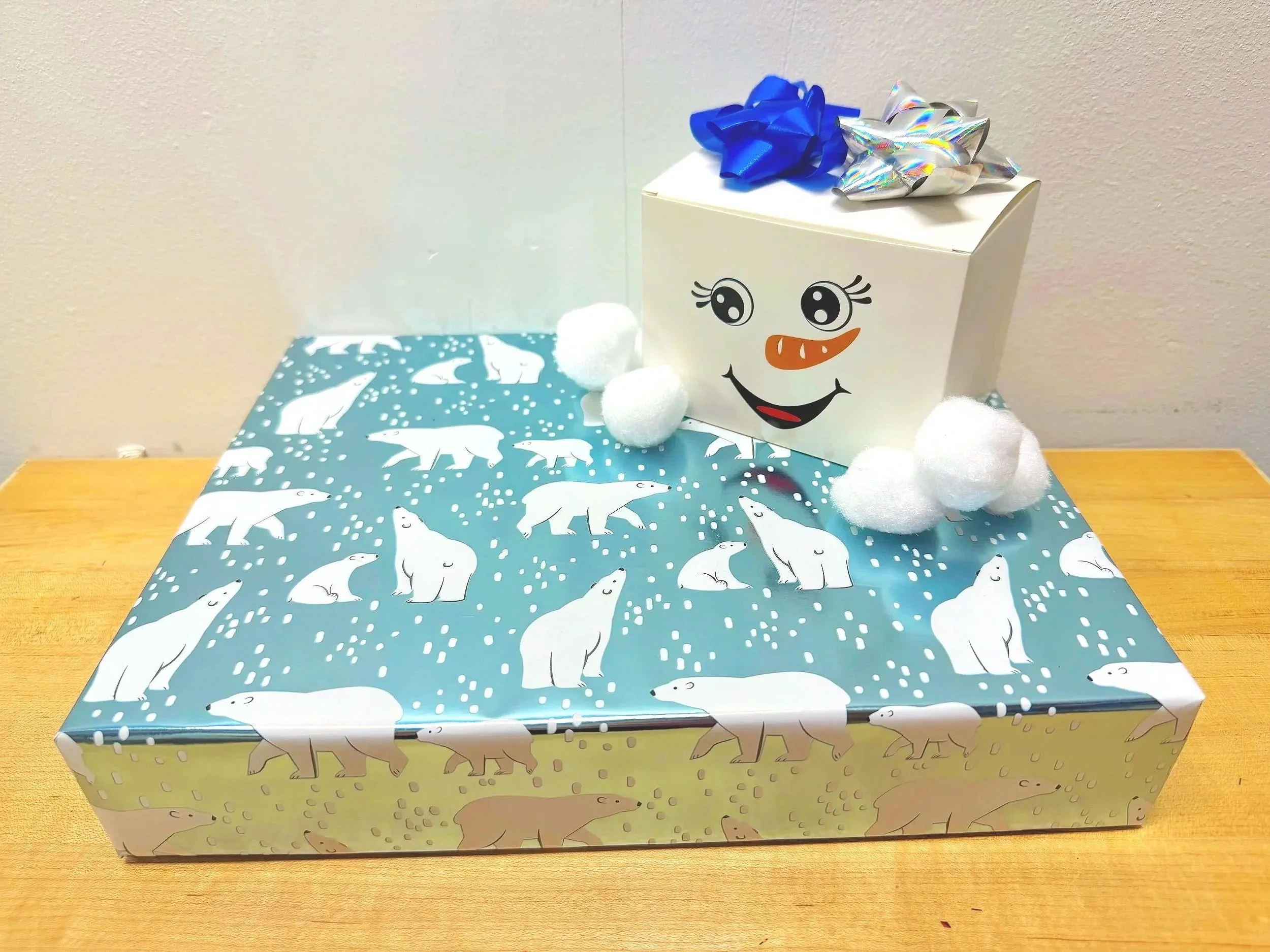 A wrapped gift with polar bears on the wrapping paper, a white box decorated with a smiling face, eyes, and built-in snowman accessories with a blue and silver bow on top, and cotton balls resembling snow on a wooden surface against a plain wall.