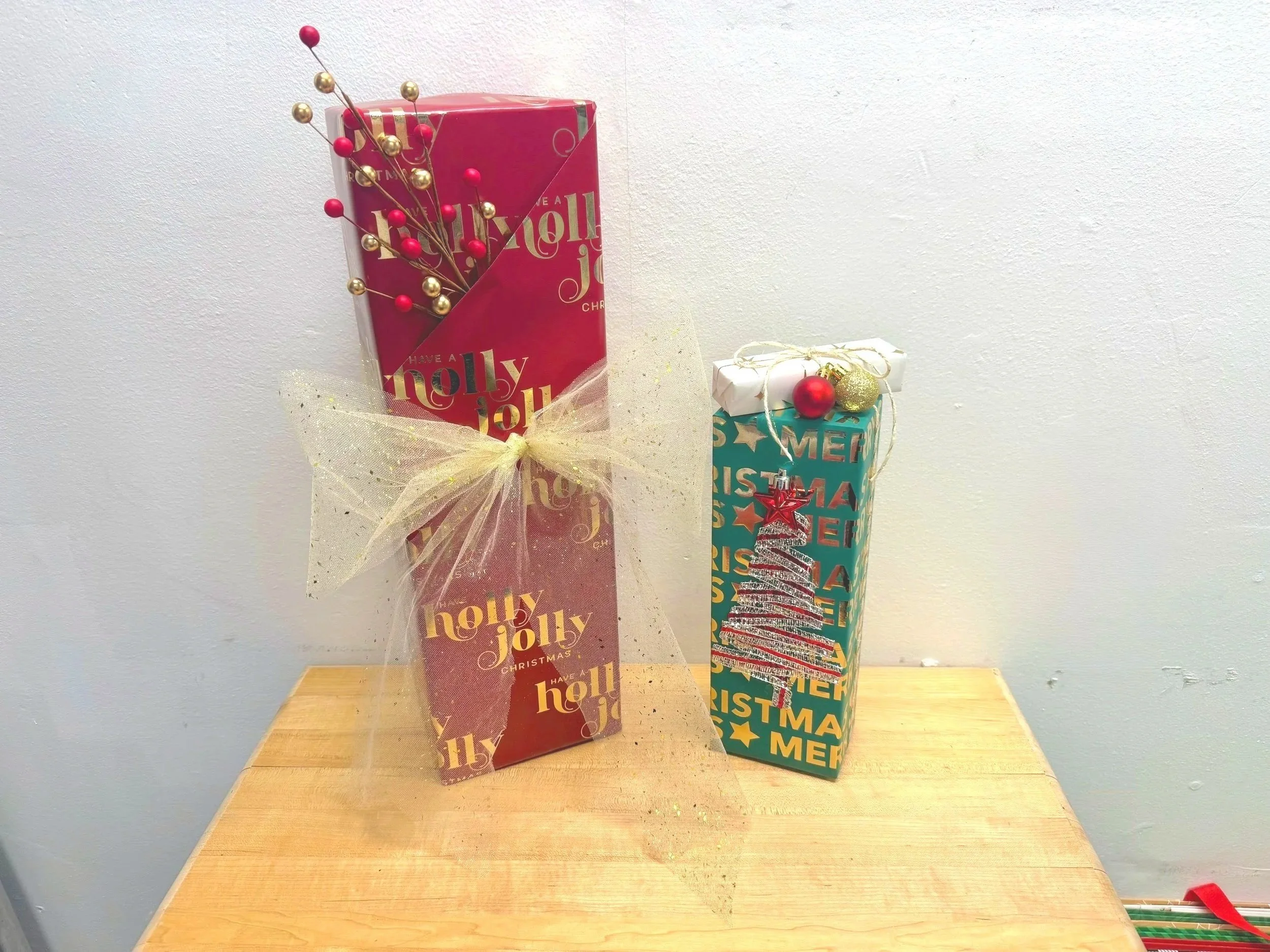 Two Christmas gift boxes on a wooden table, one wrapped in red holiday paper with gold and red decorative sticks, and the other in green paper with holiday messages, a silver ribbon, and a Christmas tree ornament.