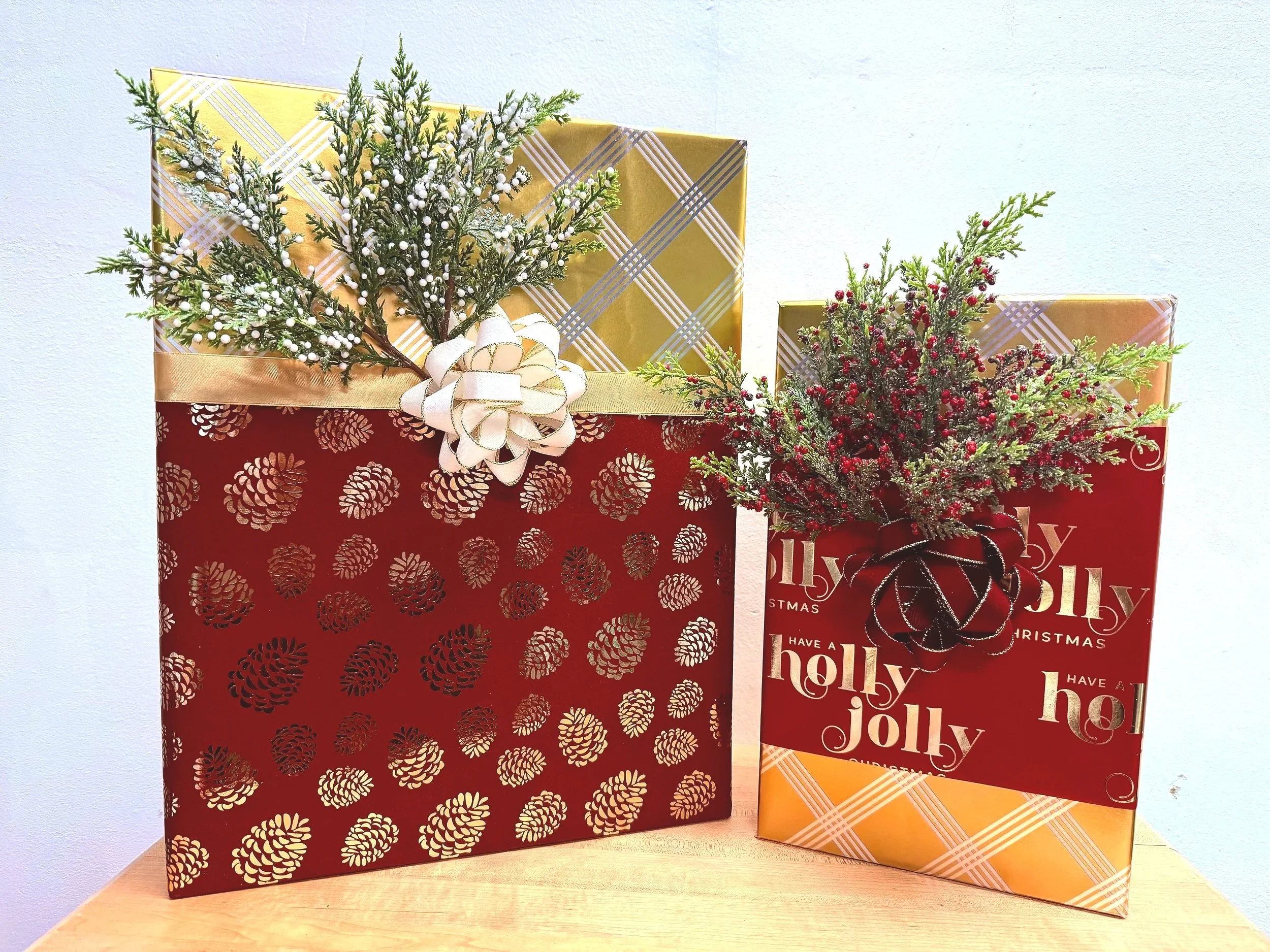 Two decorated Christmas gift boxes with greenery, one gold and one red, on a wooden surface against a light-colored wall.