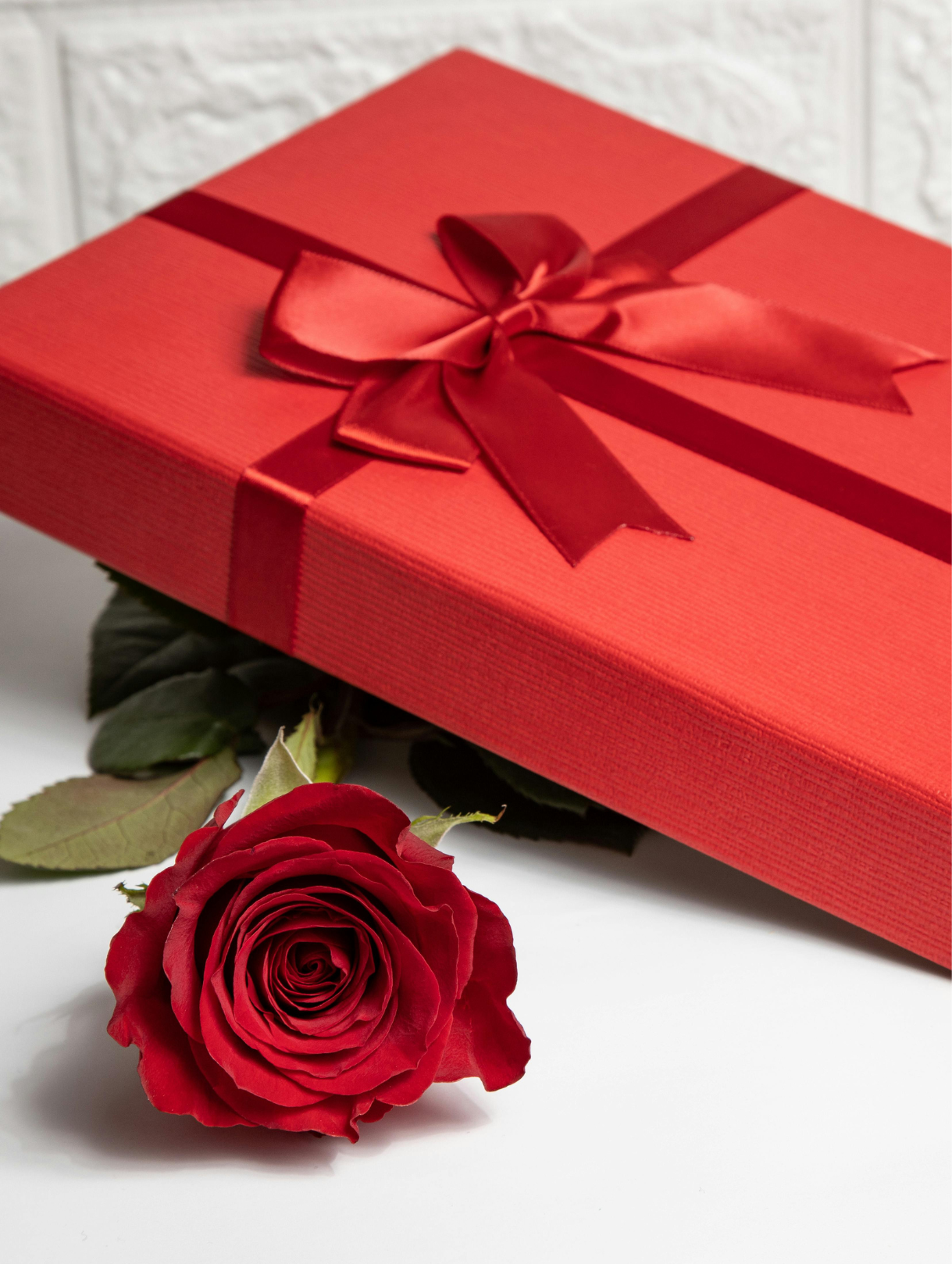 A red gift box with a shiny ribbon and bow, placed on a white surface with a single red rose next to it.