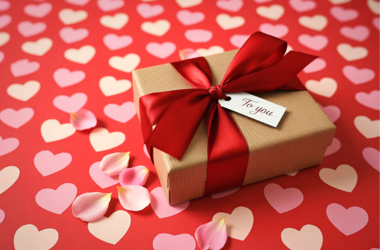 A gift box wrapped in brown paper with a red satin ribbon and bow, placed on a pink and red heart-patterned background with pink rose petals scattered around, and a white tag that reads "To you."
