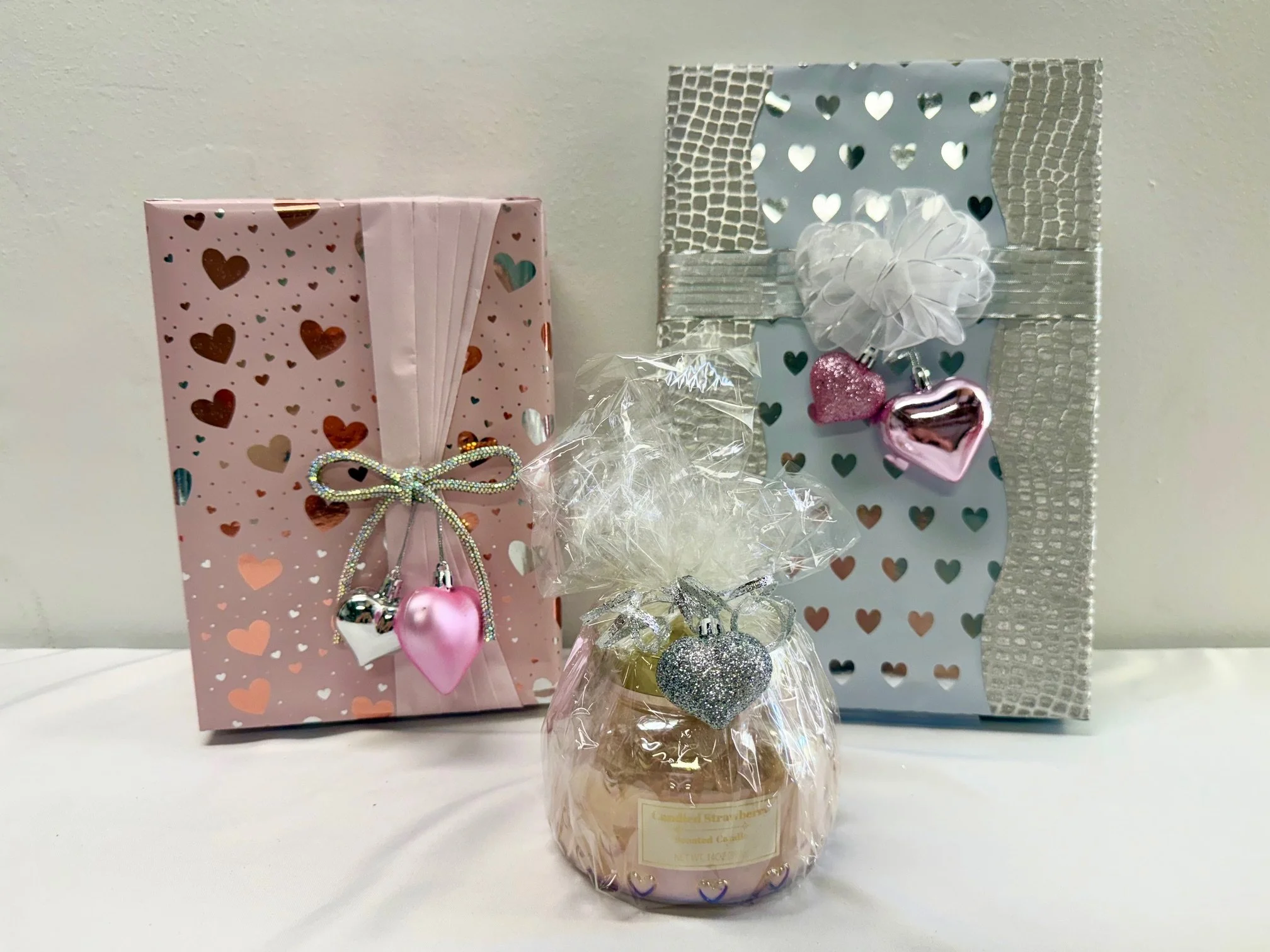 Gift bags decorated with heart patterns and charms, and a jar candle with heart and star decorations inside, all pink and silver themed.