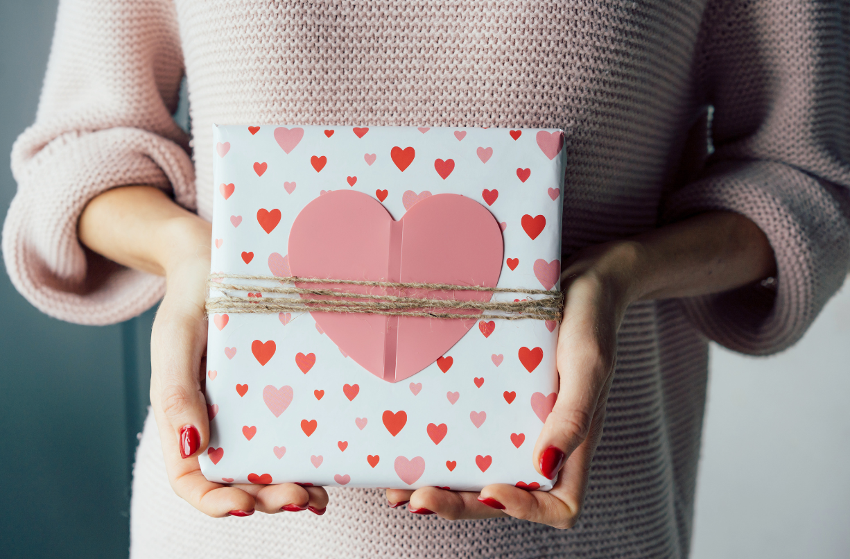 Person holding a wrapped gift box decorated with pink and red hearts, tied with twine, and featuring a large pink heart on top.