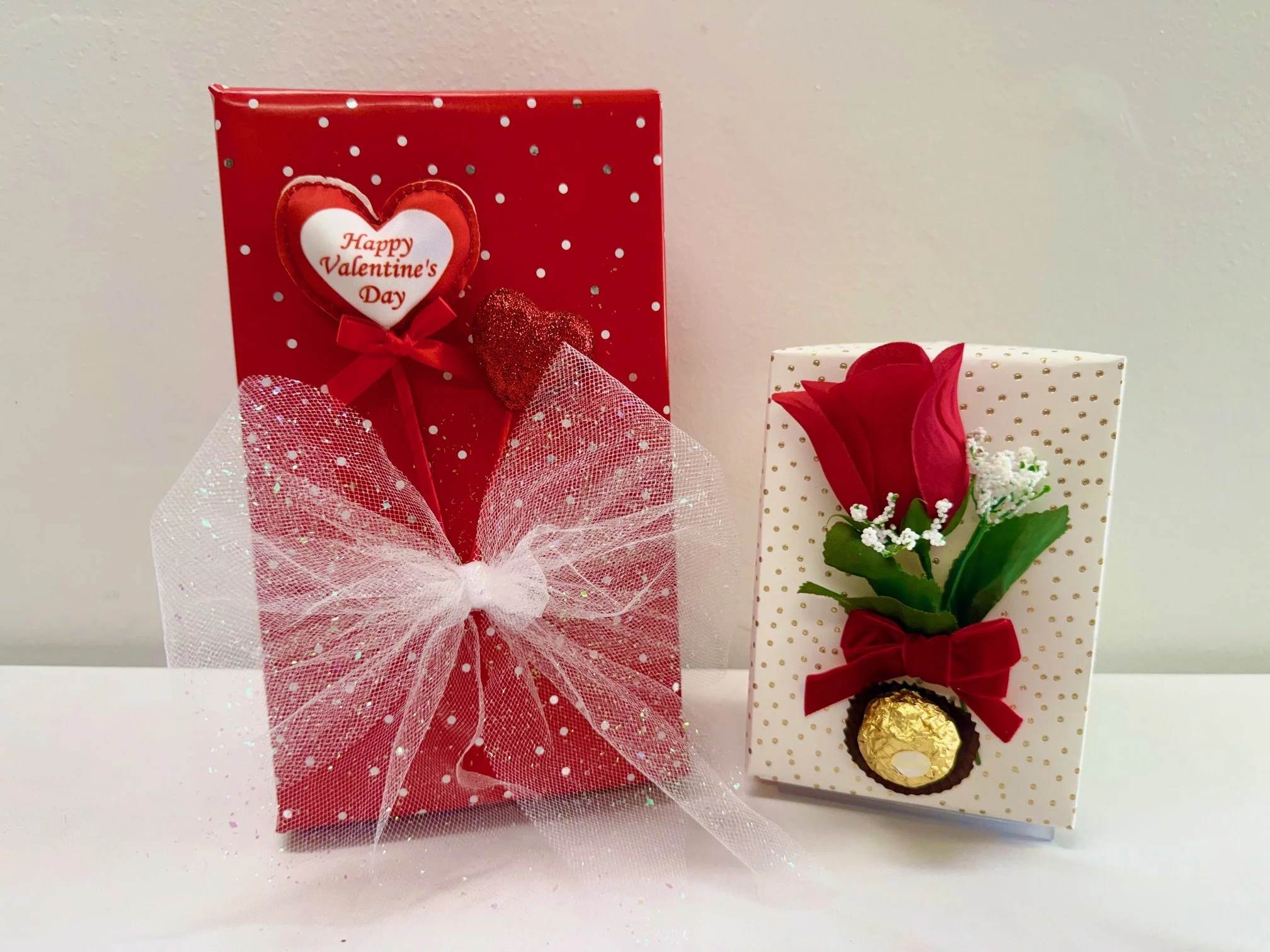 Two decorated Valentine’s Day gift boxes with flowers and chocolates, one red with a heart and ribbon, one white with a red flower and bow.