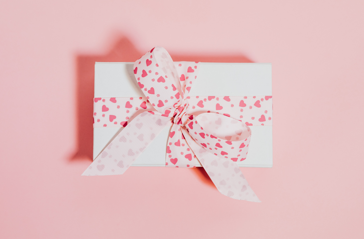 A white gift box with a pink heart-patterned ribbon and bow on a pink background.