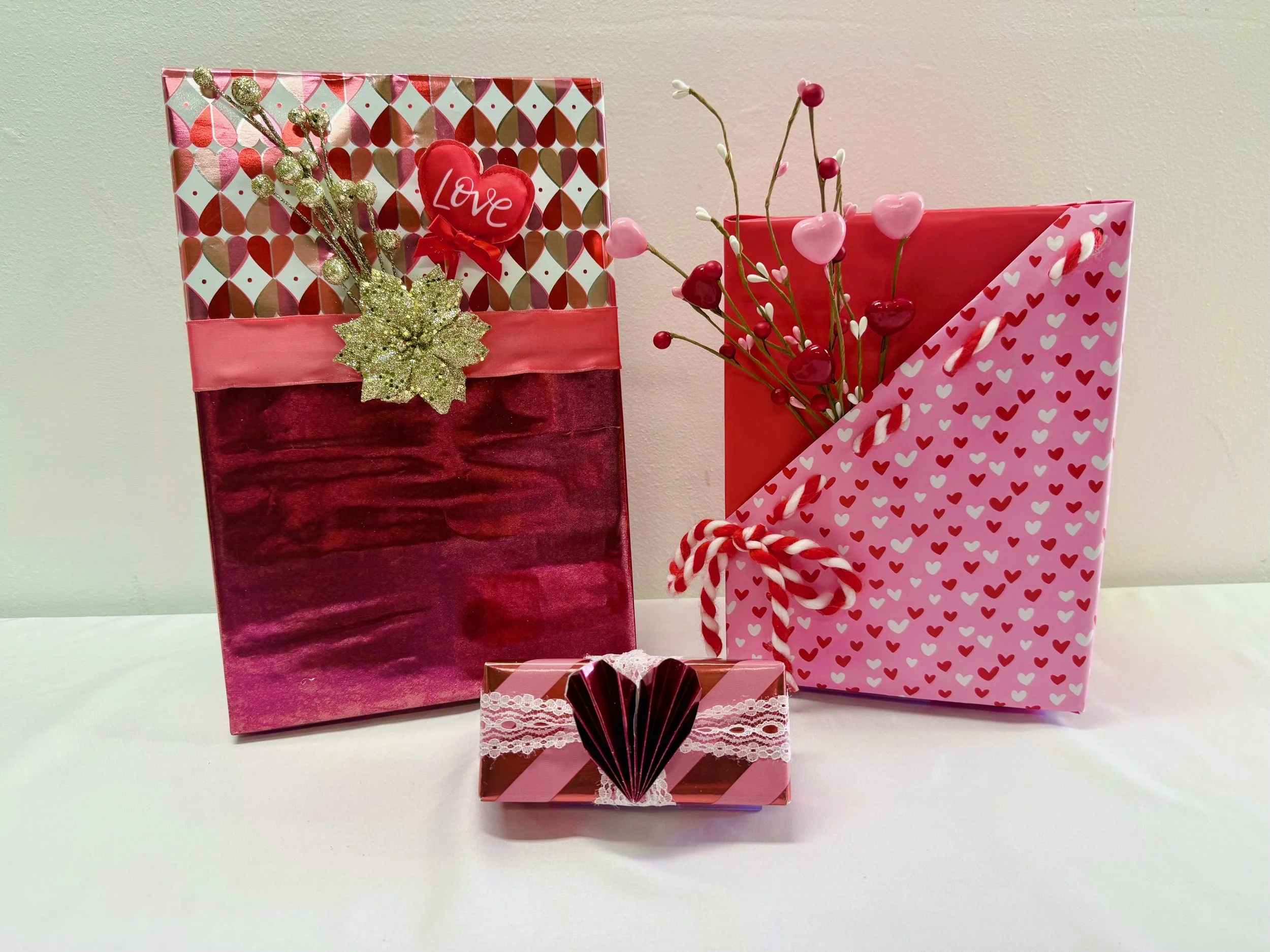 Three Valentine's Day gift bags decorated with hearts, ribbons, and flowers on a white surface against a plain wall.