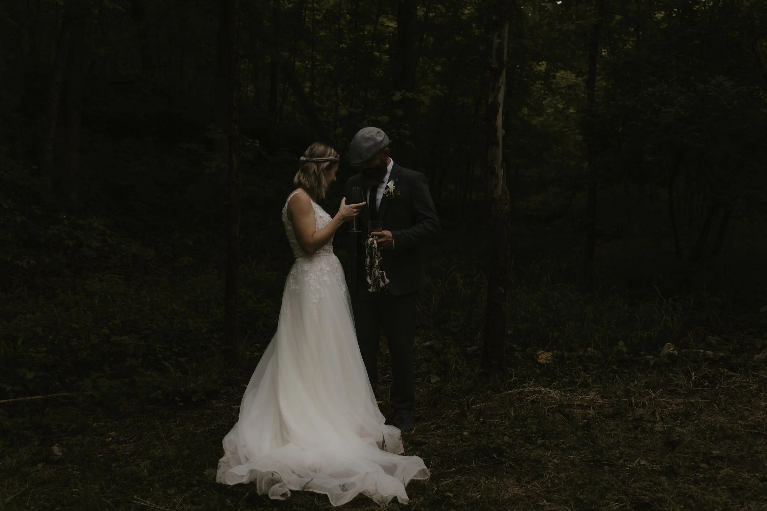 A bride in a white wedding gown and a groom in a dark suit with a hat holding glasses in a forest, standing close together.