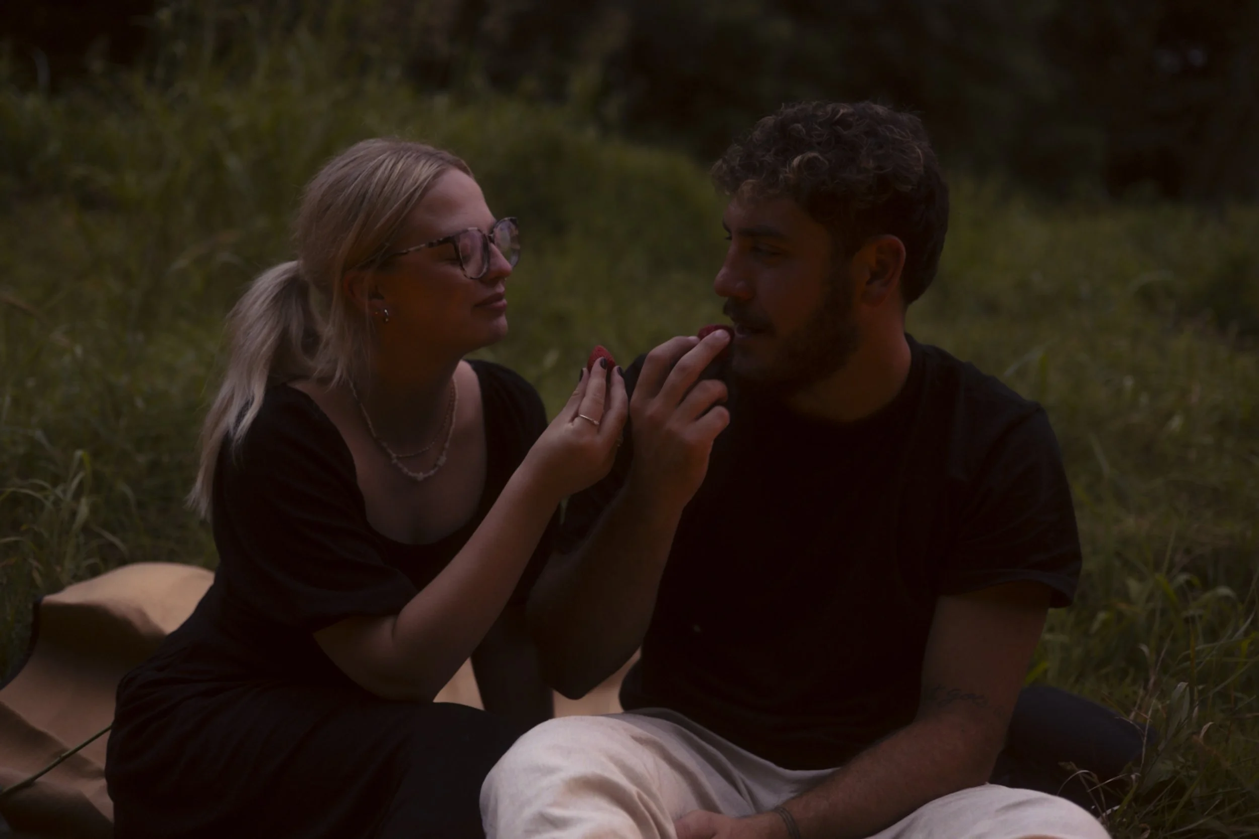 A woman and a man sitting outdoors on grass during dusk, with the woman offering the man a snack.