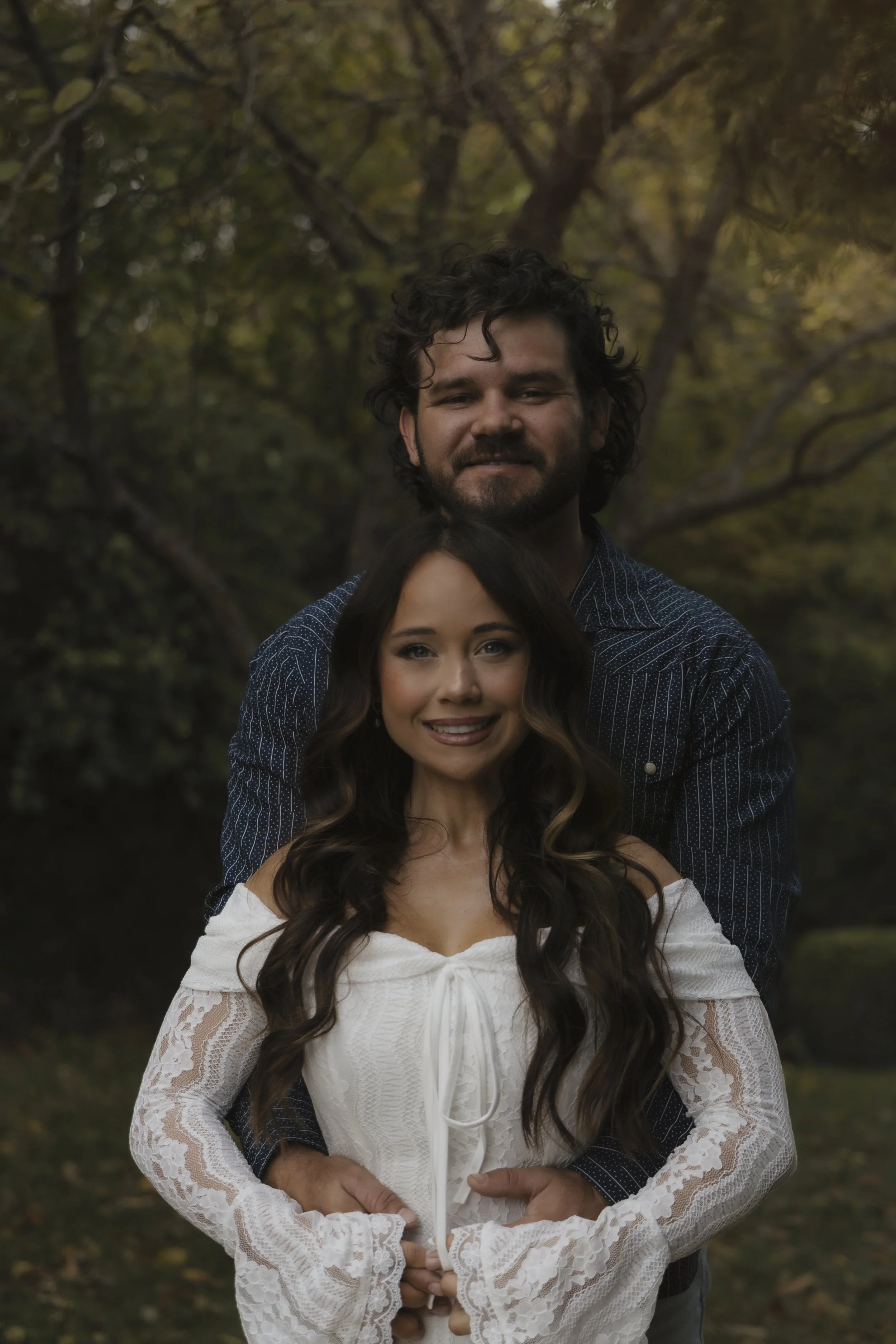 A couple standing outdoors in front of trees, with the man standing behind the woman, both smiling.