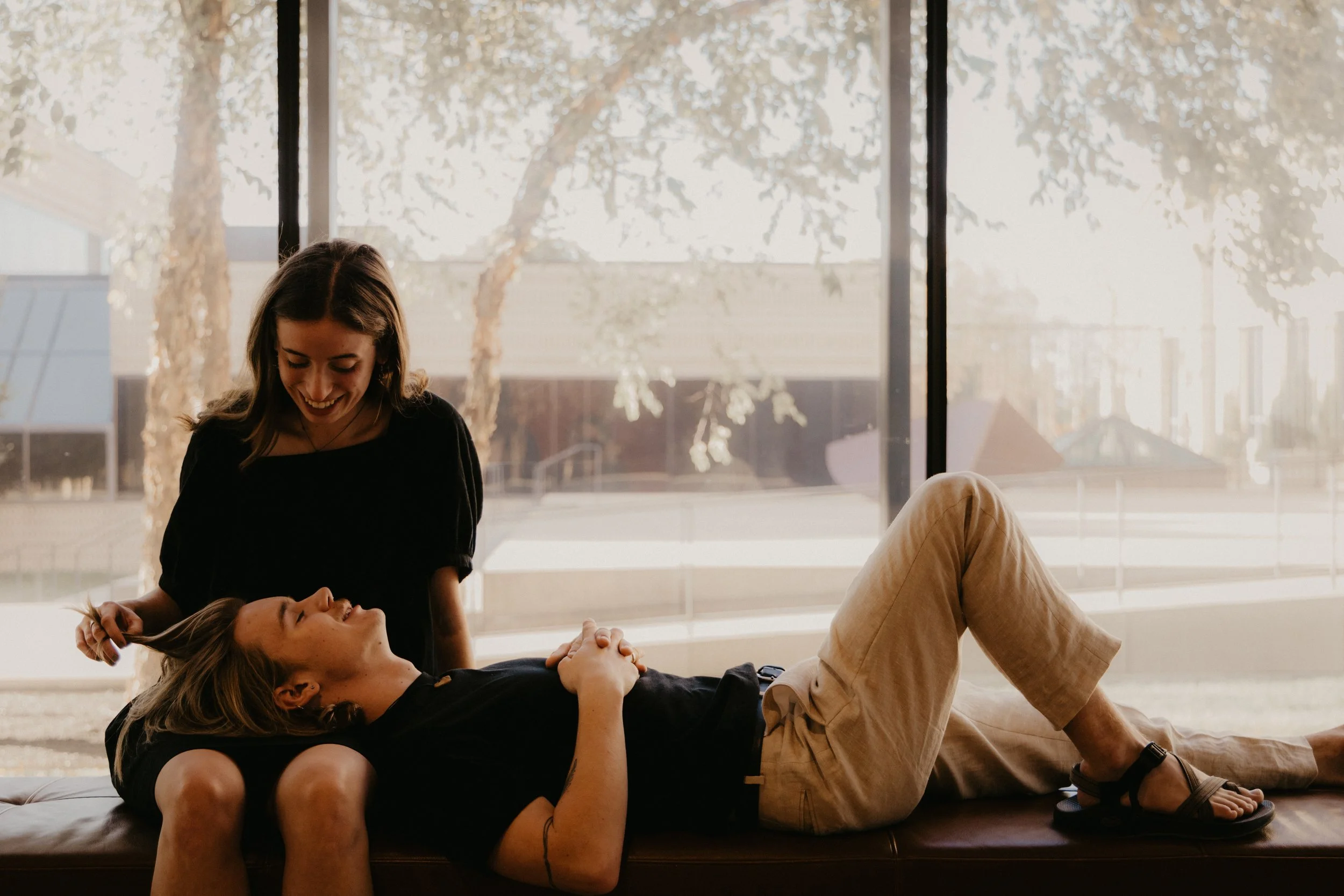 Two women smiling and engaging in a playful moment indoors near a large window with trees outside.
