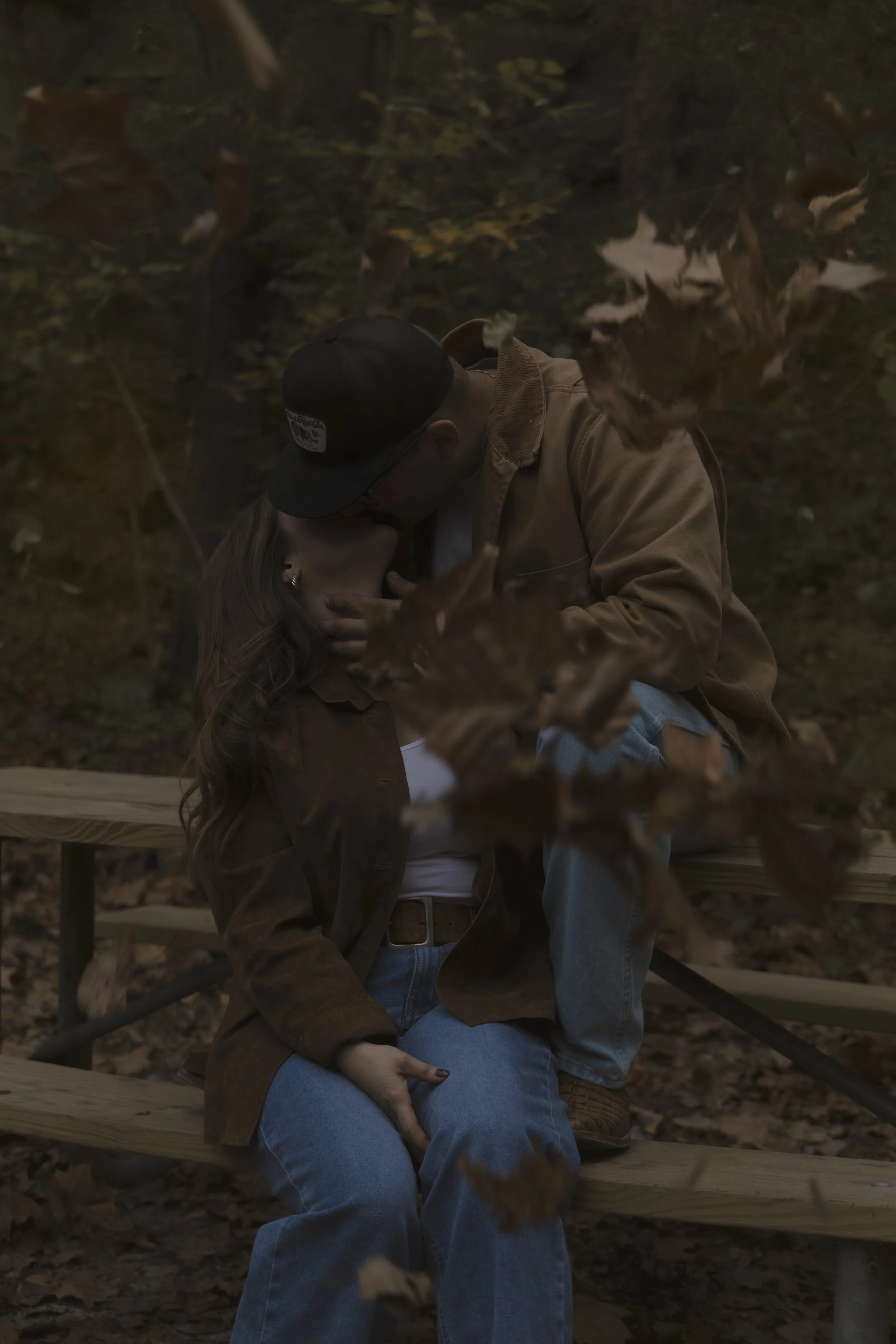 A romantic couple sharing a kiss, sitting on park benches surrounded by falling autumn leaves.