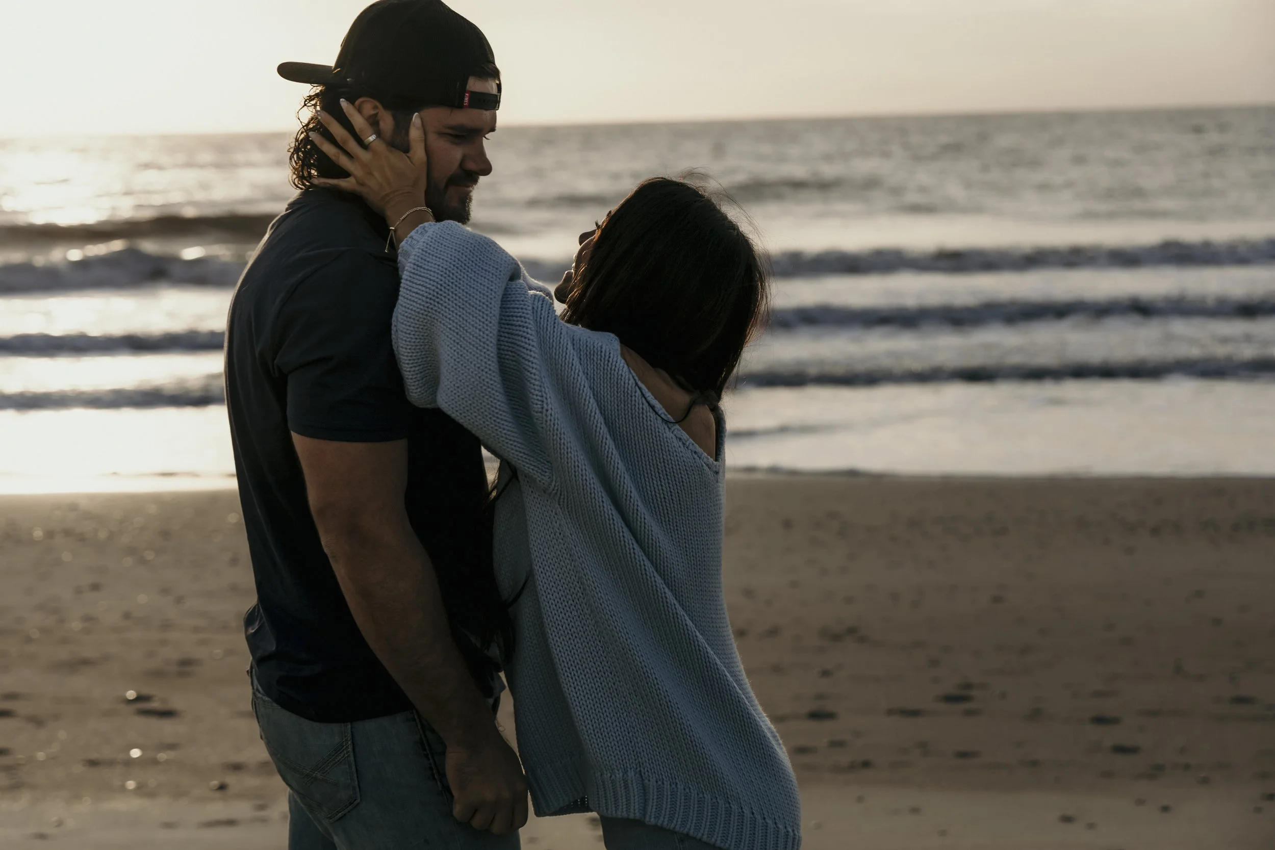 A couple embracing on a beach at sunset, with the man holding the woman's face and the woman touching the man's face, both smiling softly.