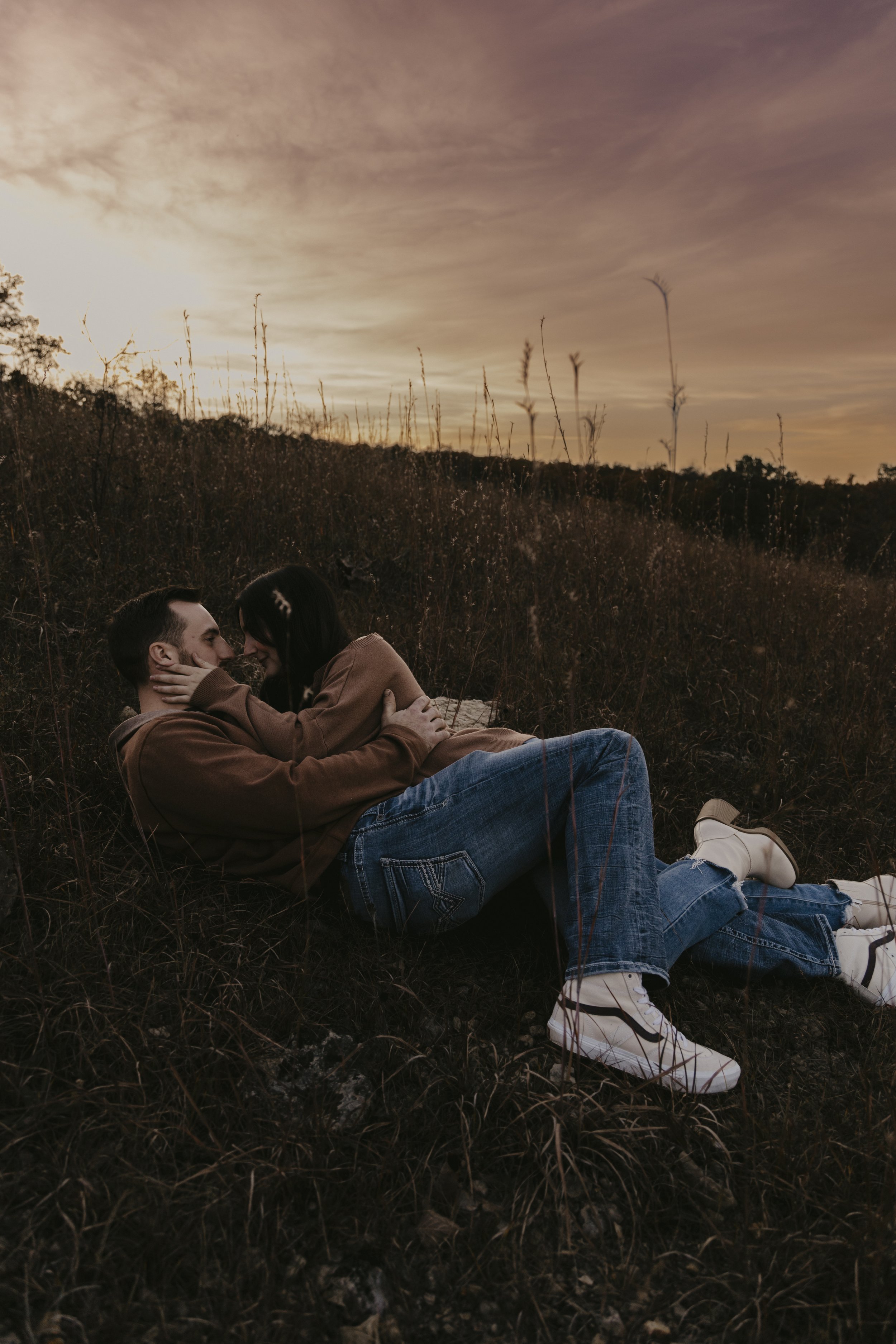 A couple lying on the ground in a field at sunset, embracing and looking at each other.