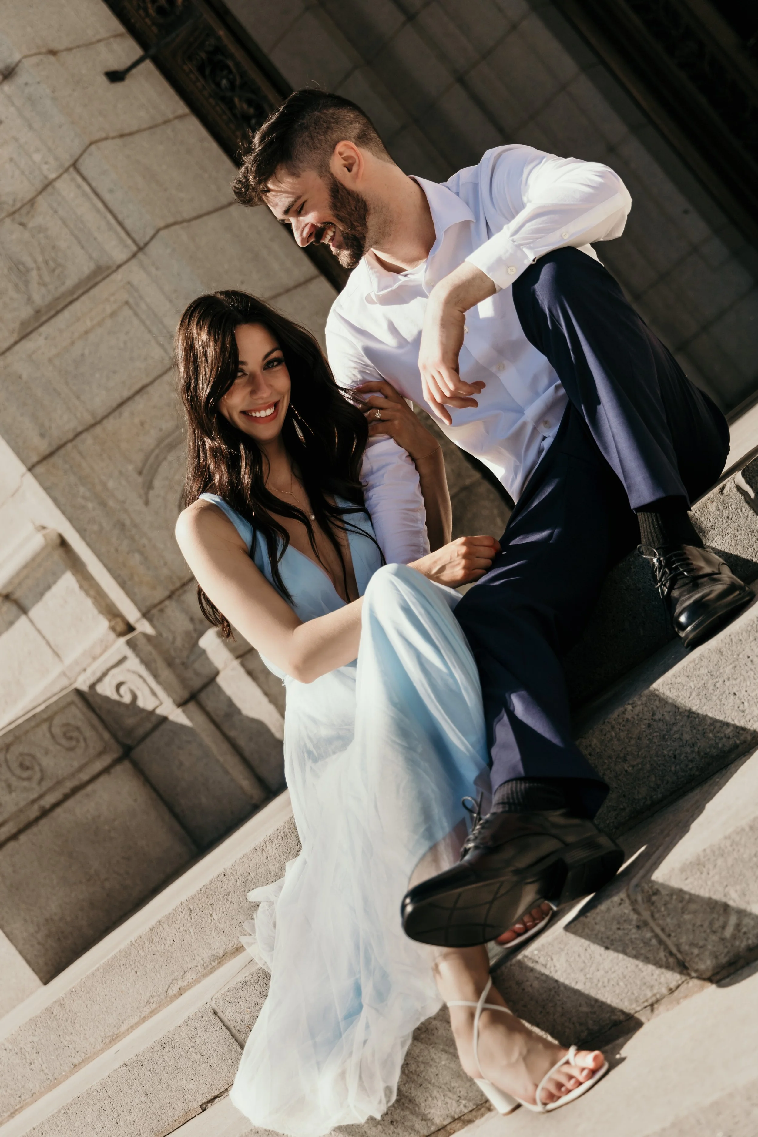 A young man and woman sitting on a stone ledge outside a building, laughing and smiling at each other. The woman is wearing a light blue dress and high heels, and the man is dressed in a white shirt and dark pants.