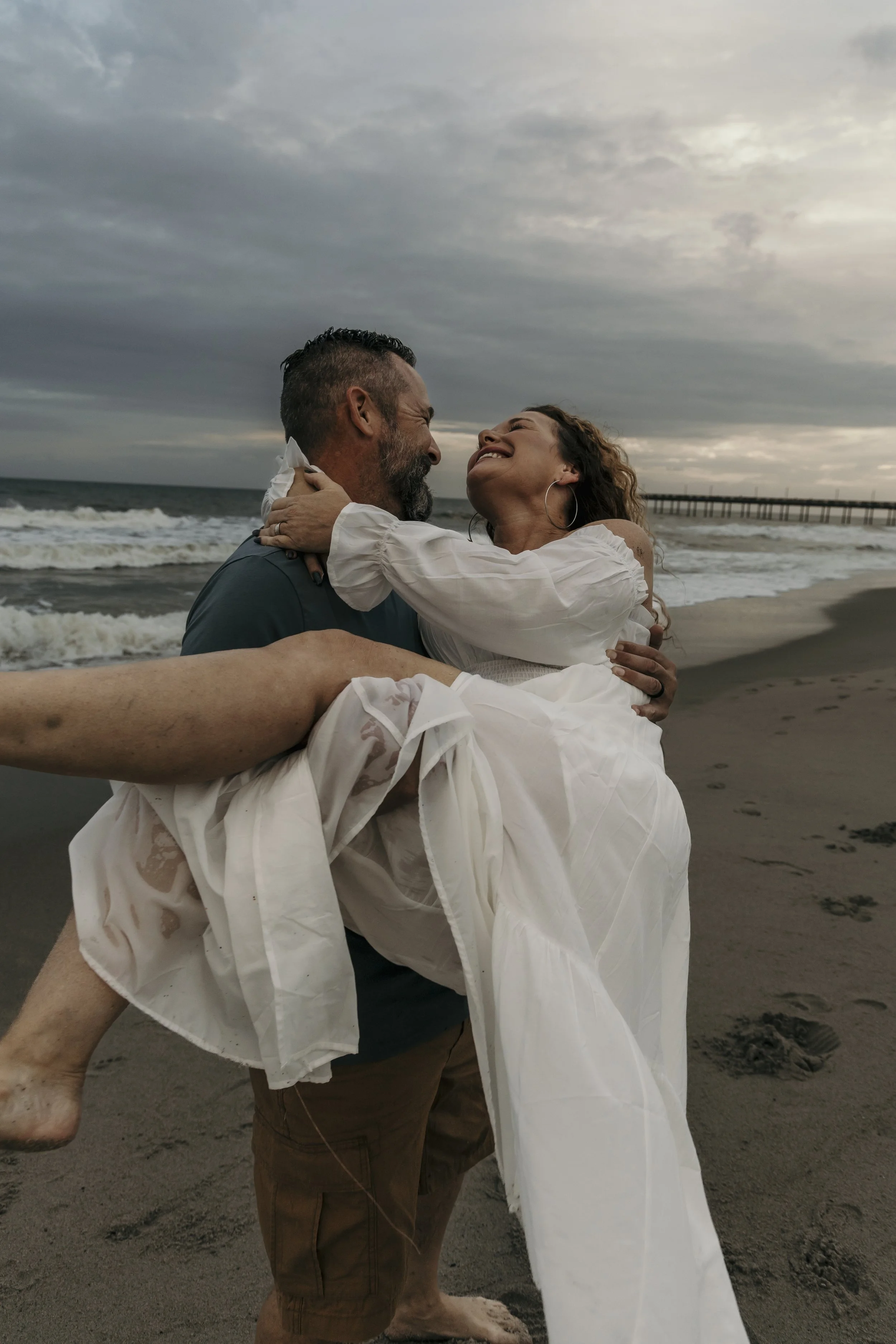 A man holding a woman in a white dress at the beach, both smiling and looking at each other, with ocean waves and a cloudy sky in the background.