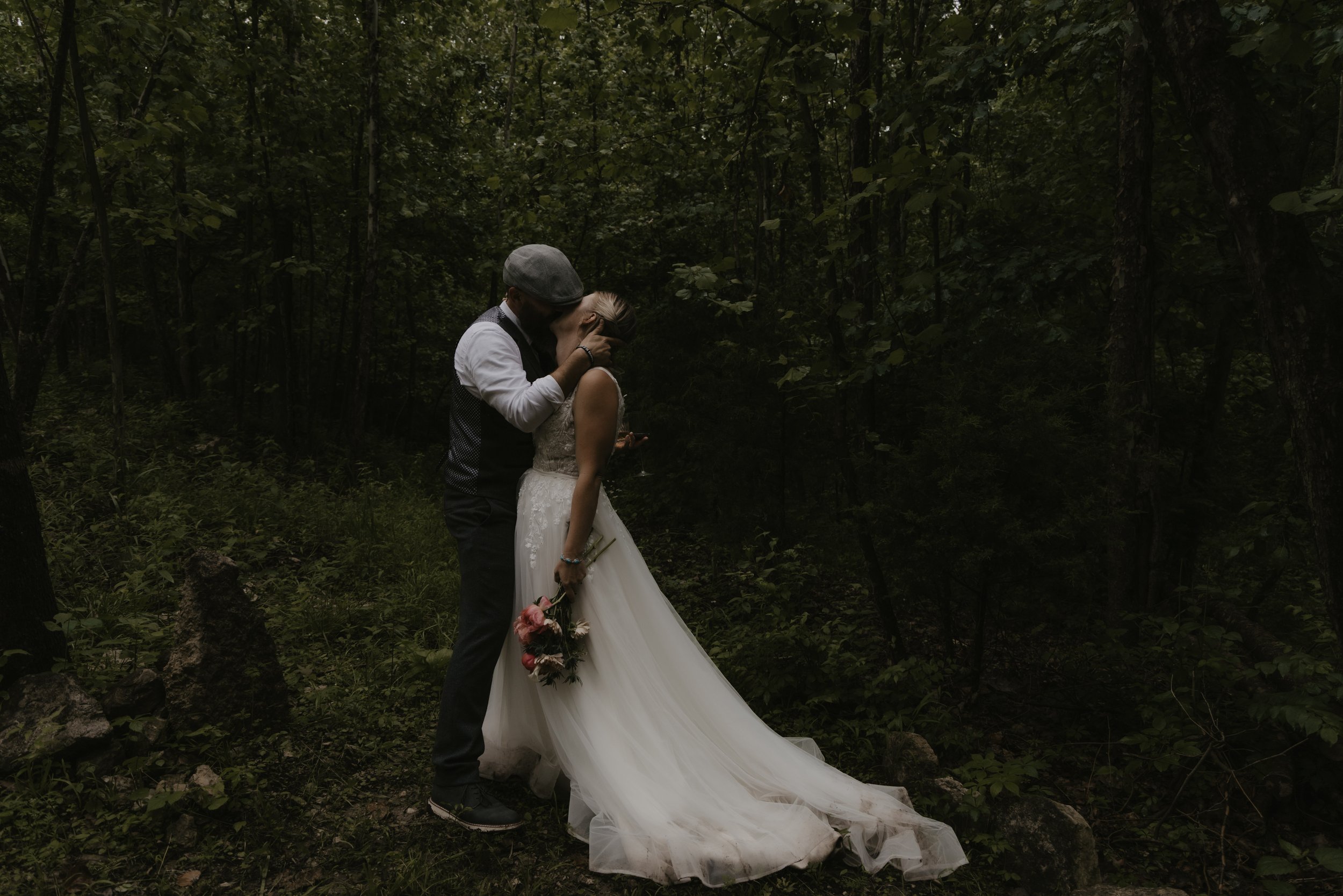 A couple dressed in wedding attire sharing a kiss in a lush, green forest environment. The bride is holding a bouquet of pink and white flowers.