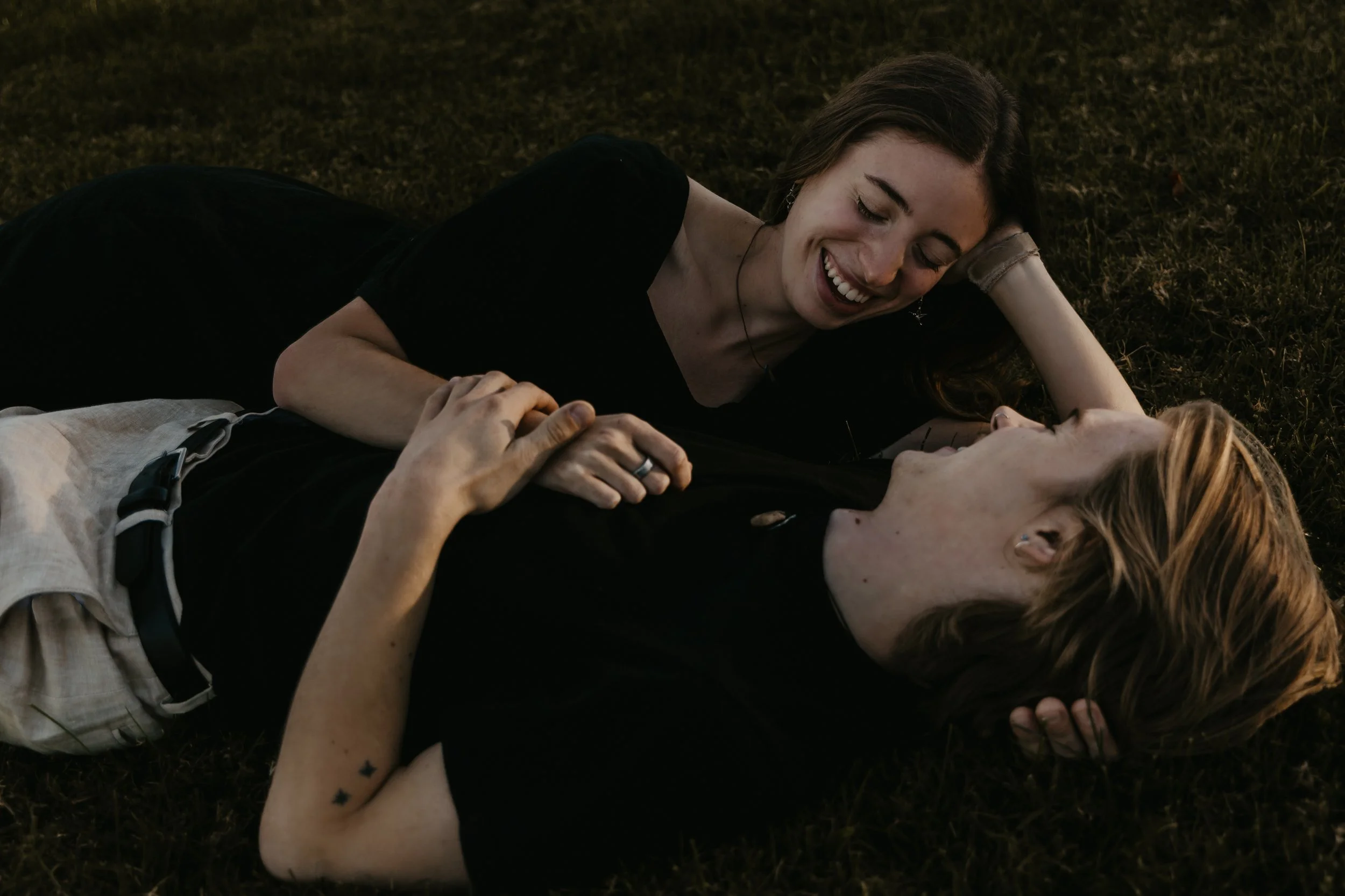 Two women lying on grass, smiling and looking at each other, with one gently touching the other's chest.