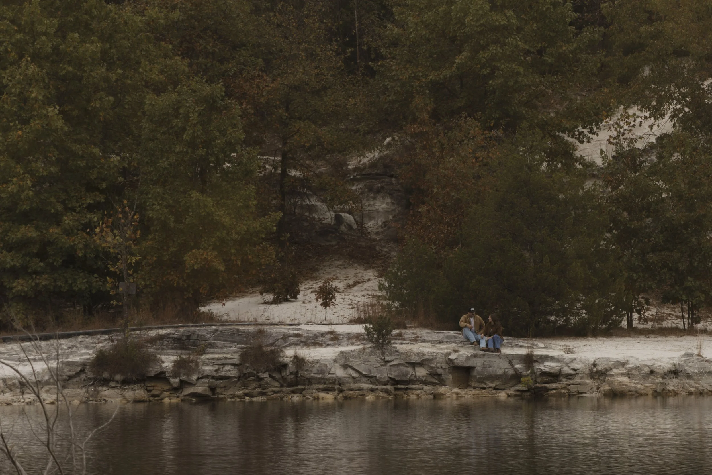 Two people sitting on a rocky riverbank surrounded by trees with fall foliage.