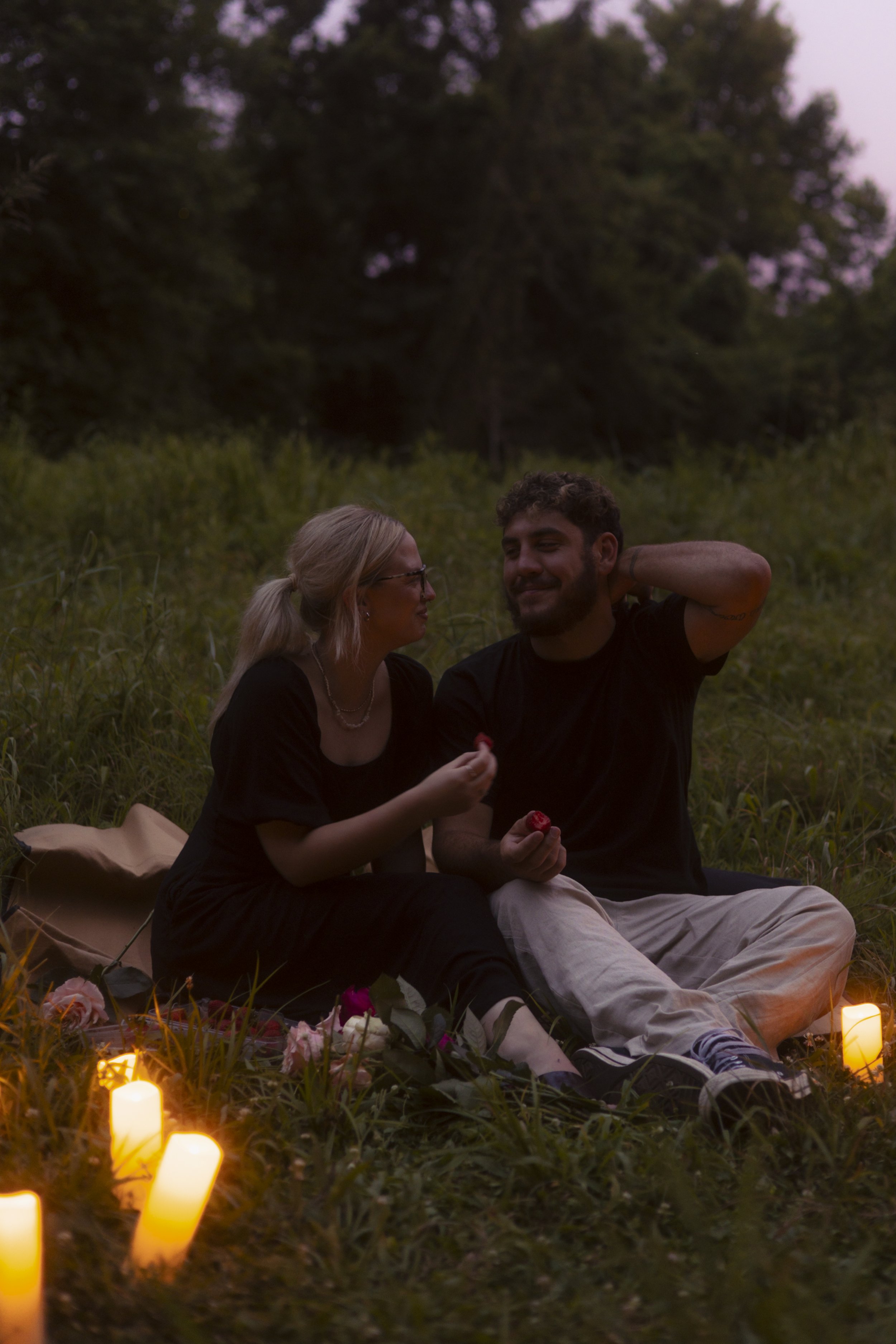 A couple having a romantic picnic outdoors at sunset, sitting on a blanket with candles and flowers around them.